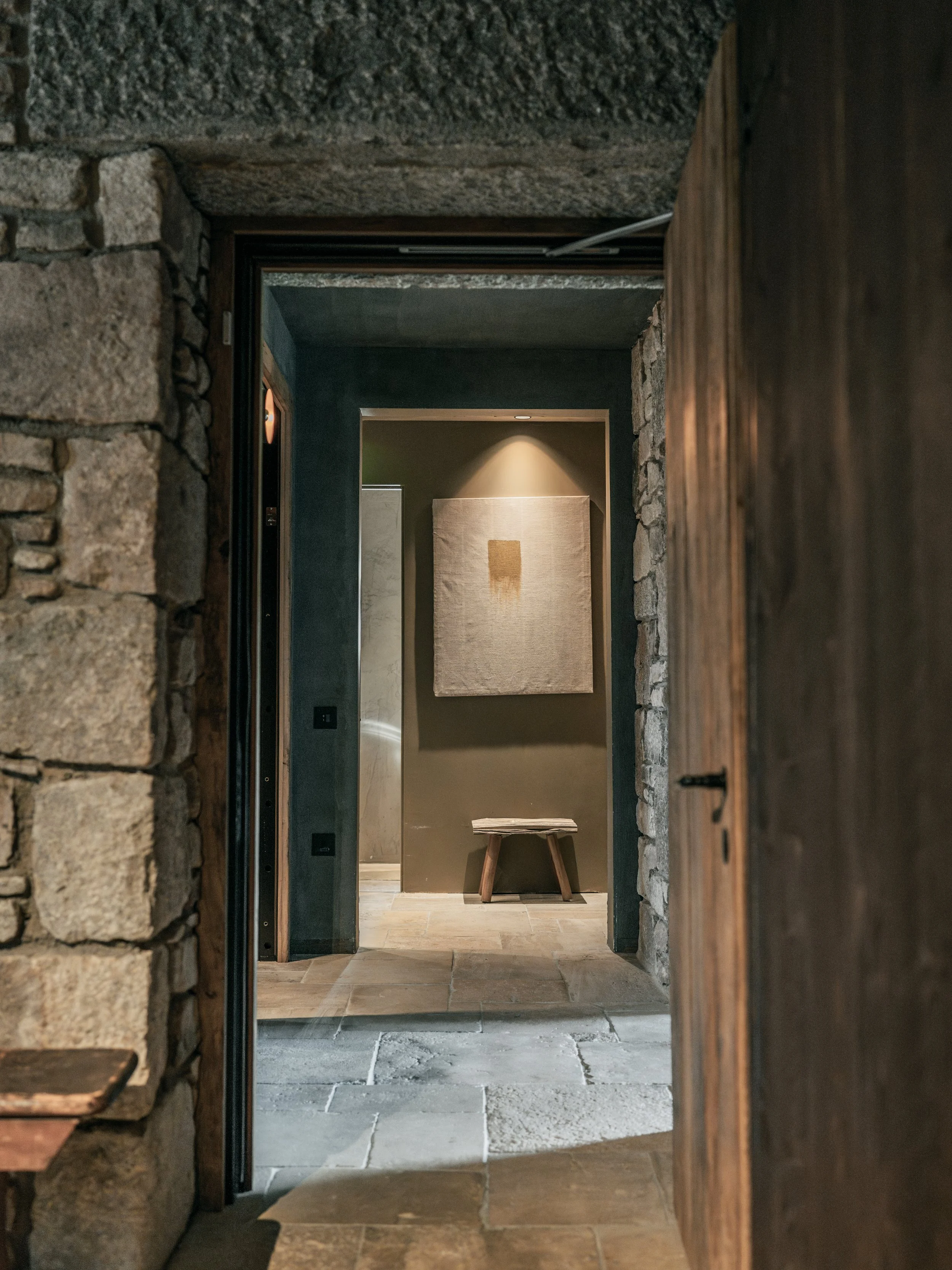 A hallway with stone walls on both sides, leading to a small room with a single wooden stool, a large beige fabric wall hanging, and a spotlight casting a glow on the wall.
