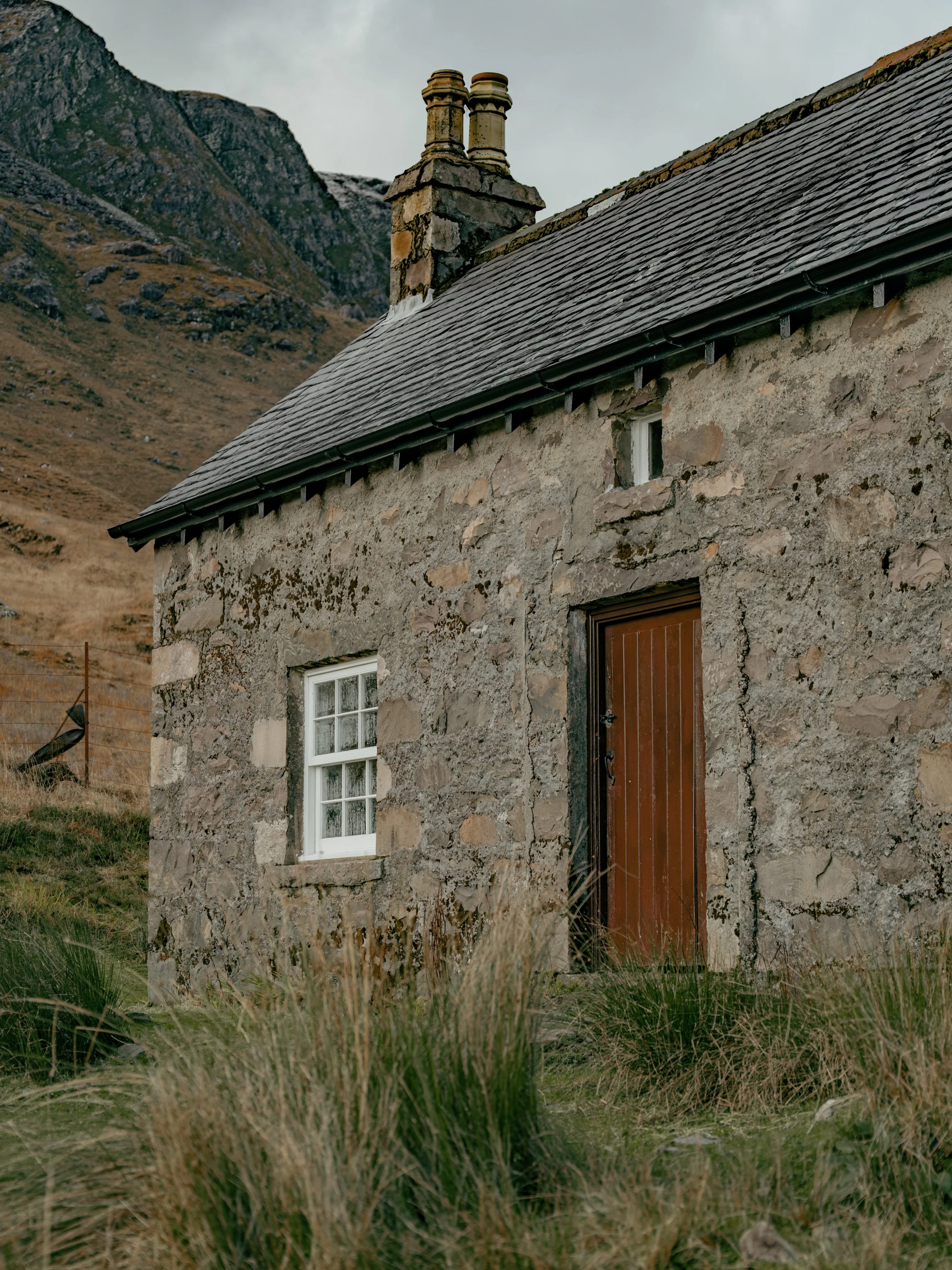 A rustic stone house with a wooden front door, small window, and stone chimney, set against a mountain landscape with tall grass in the foreground.