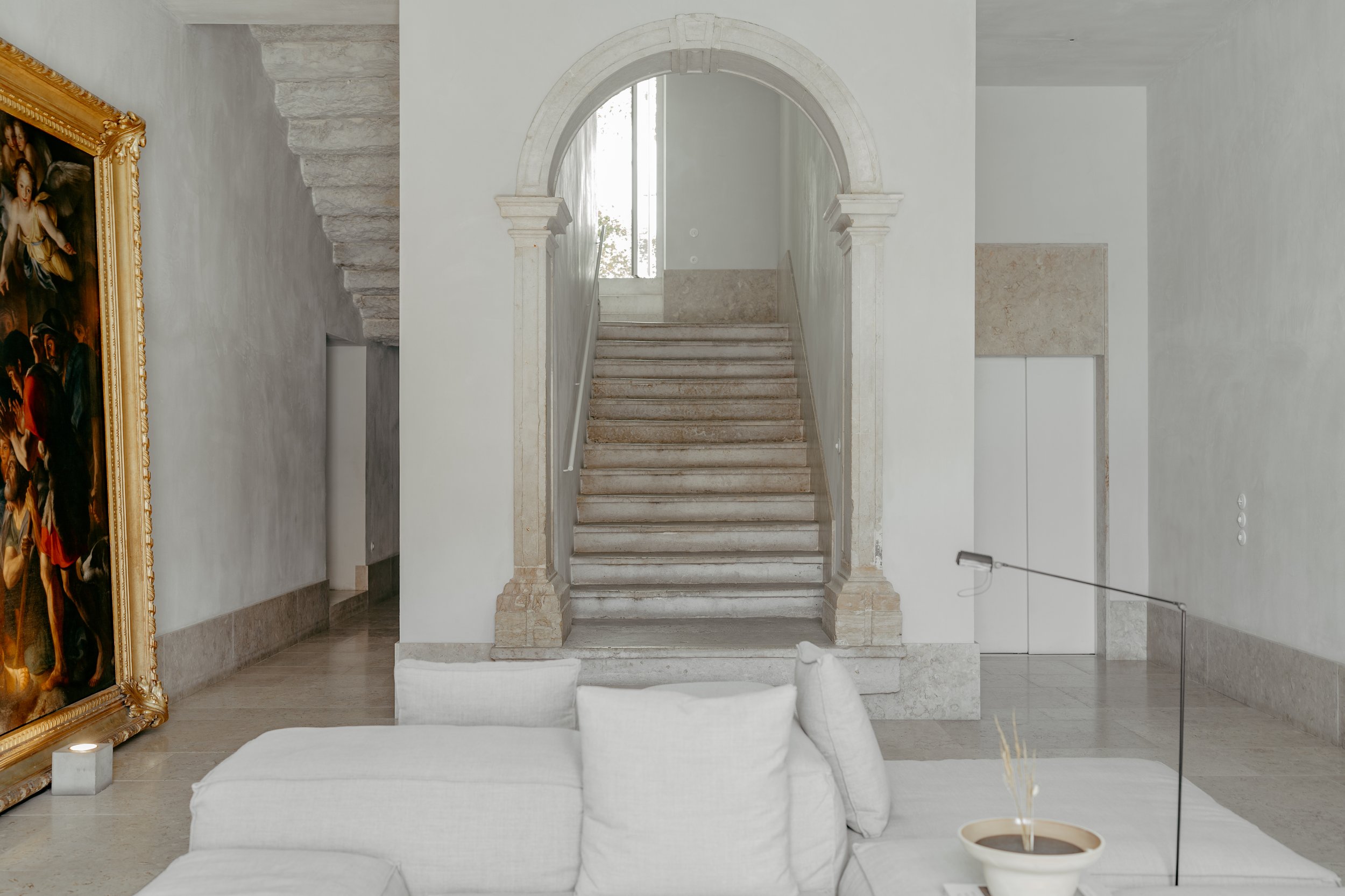 Interior of a modern house with a staircase, white walls, and a large framed painting on the left. There is a white sofa in the foreground, and a small round table with a dry plant in a pot on it. The staircase has beige marble steps and is framed by