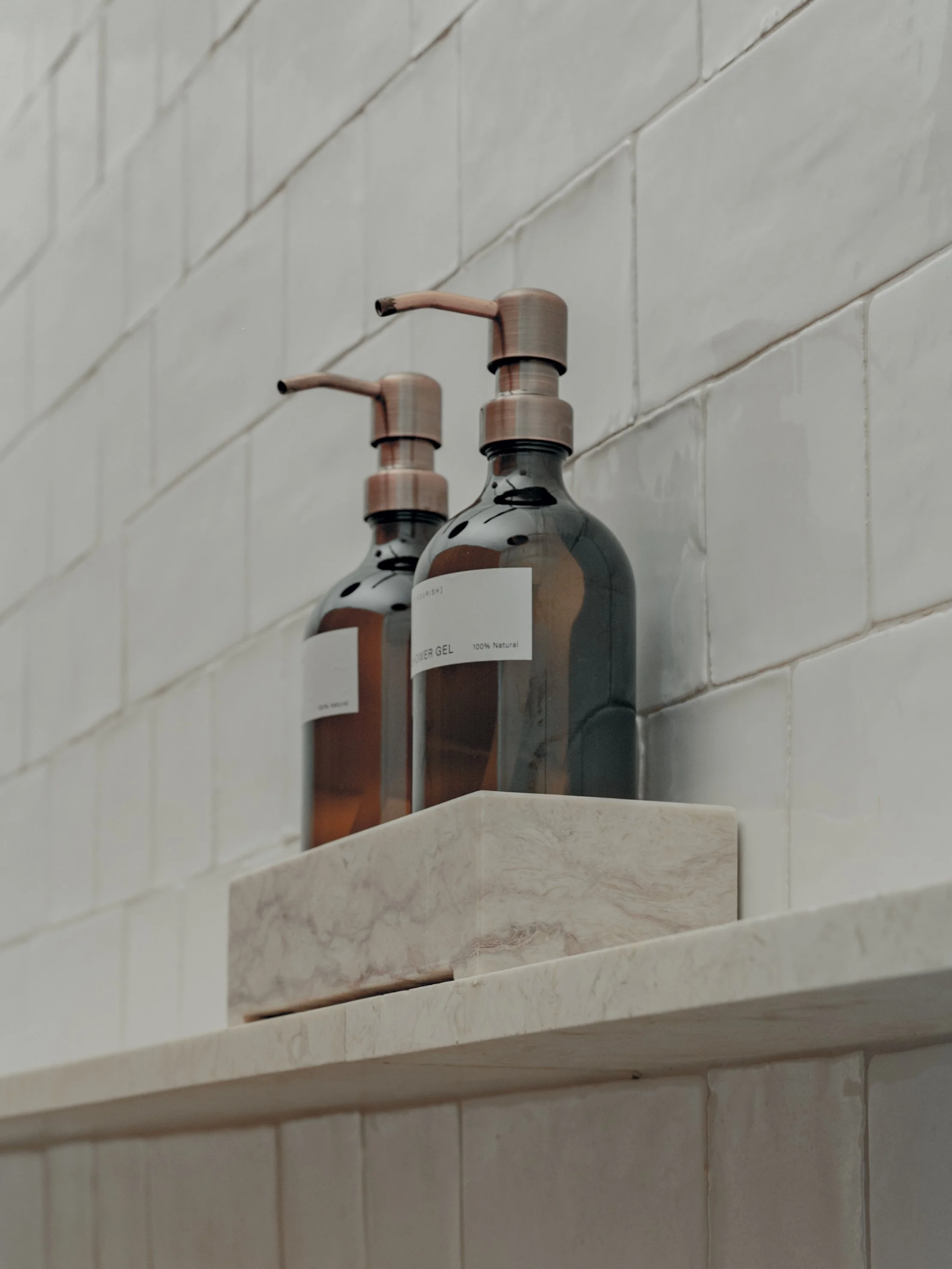 Two brown liquid soap or lotion dispensers mounted on a white tiled wall above a marble shelf.