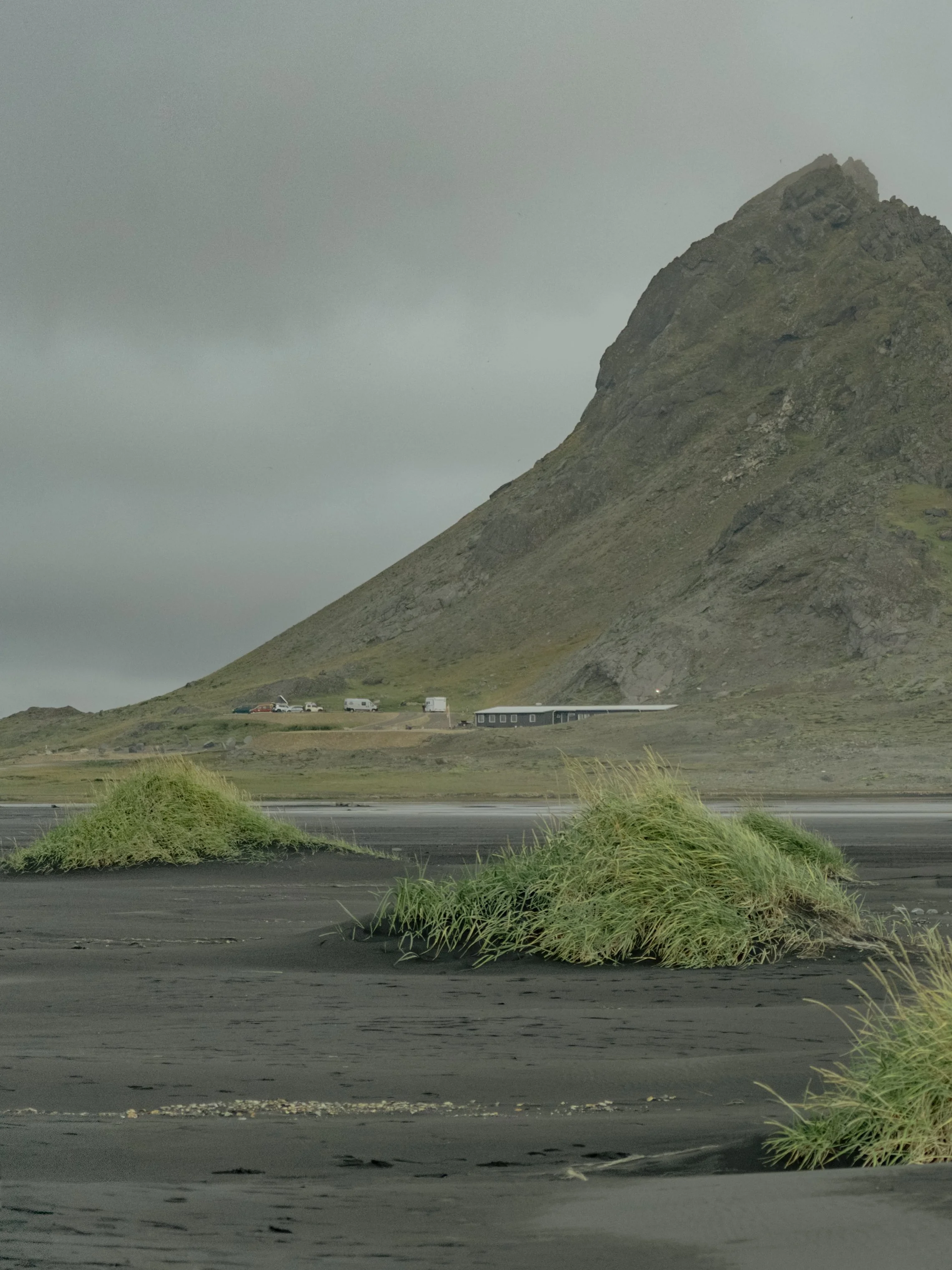 A mountain with a small building and parked cars at its base, grassy terrain, cloudy sky.