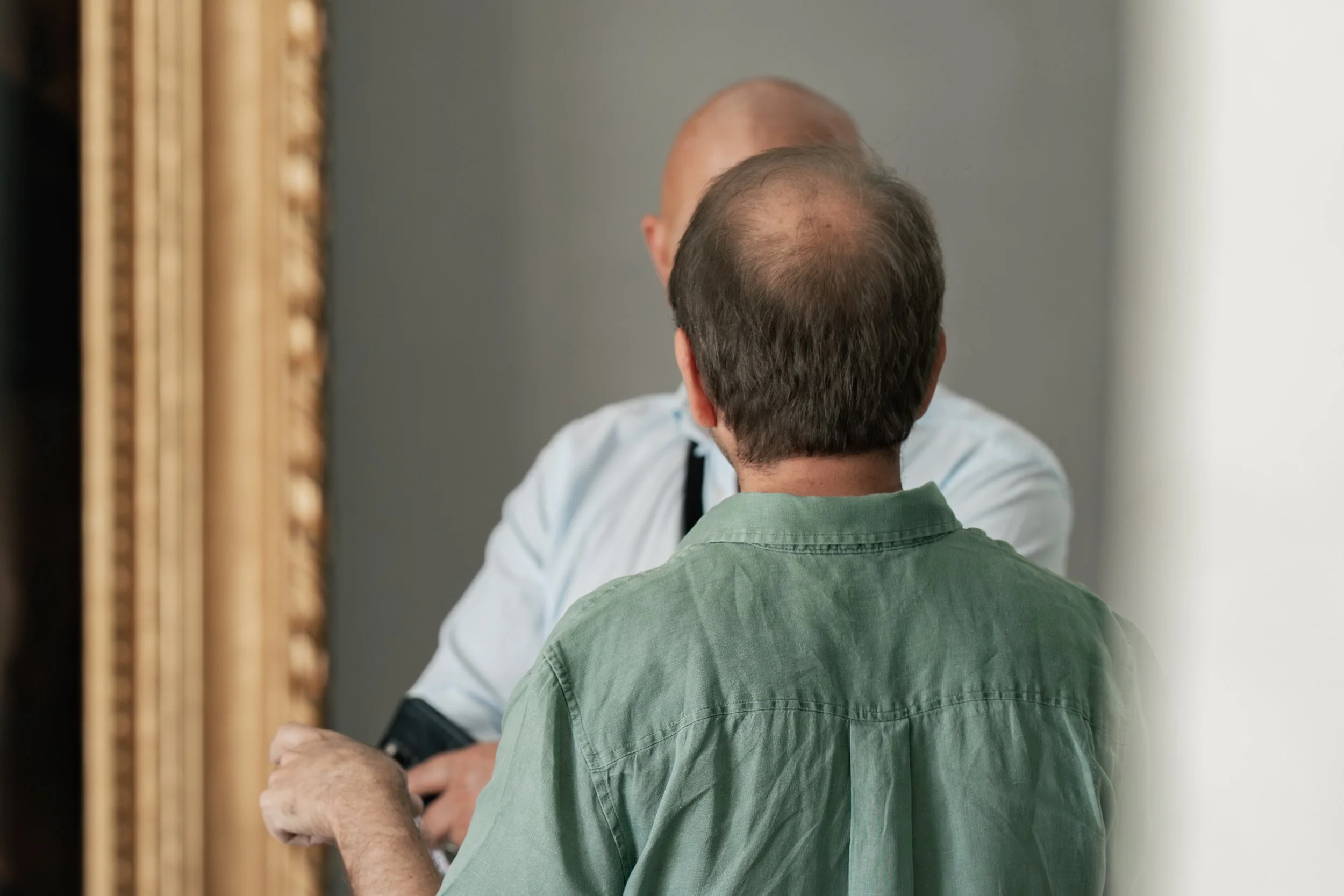 Two men are having a conversation, with one facing away from the camera and the other reflected in a mirror, revealing his face and bald head.