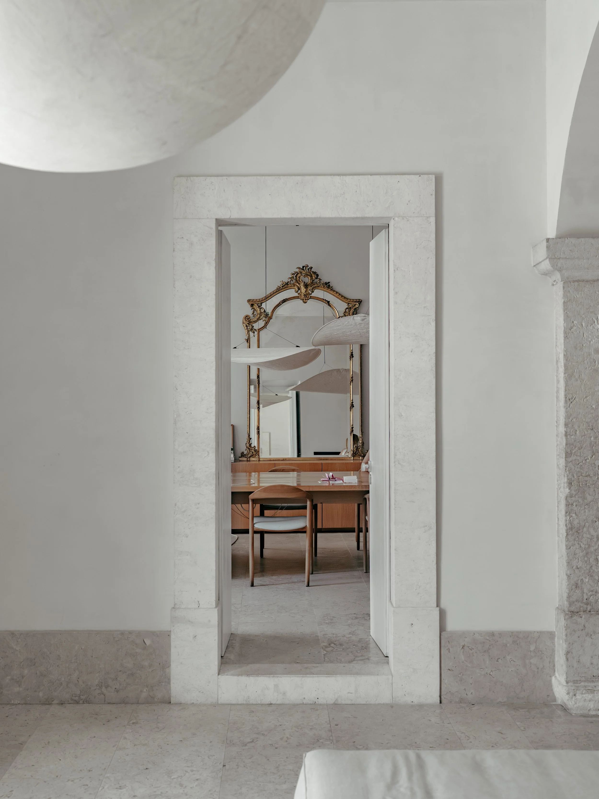 White textured walls and floor in a minimalist room, with a view through a doorway showing a wooden desk with a chair, a large ornate gold-framed mirror, and hanging fabric lights.