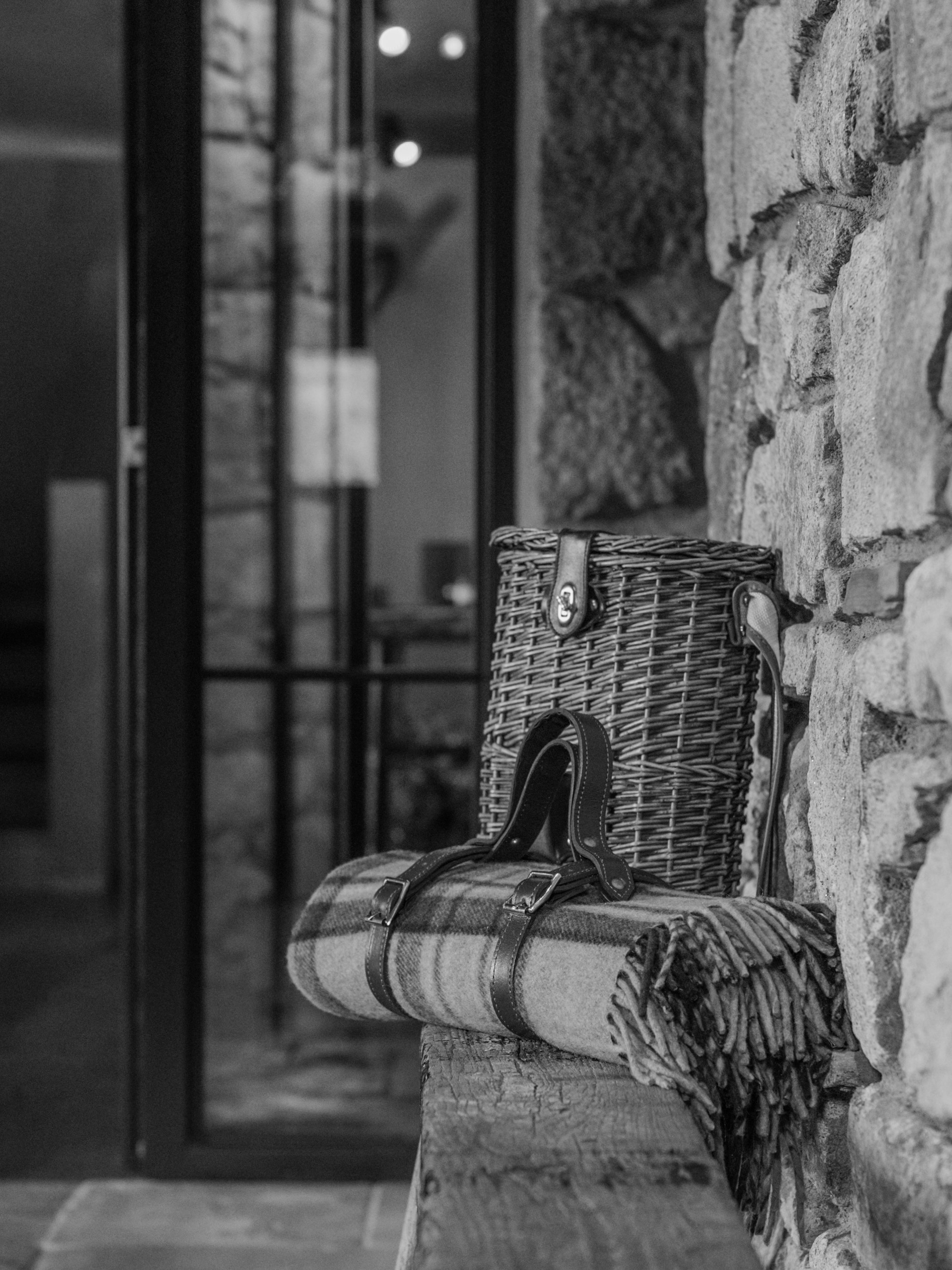 A black and white photo showing a wicker basket and a plaid blanket or scarf with leather straps on a wooden bench against a stone wall. In the background, there is a window door with reflections and a visible interior space.