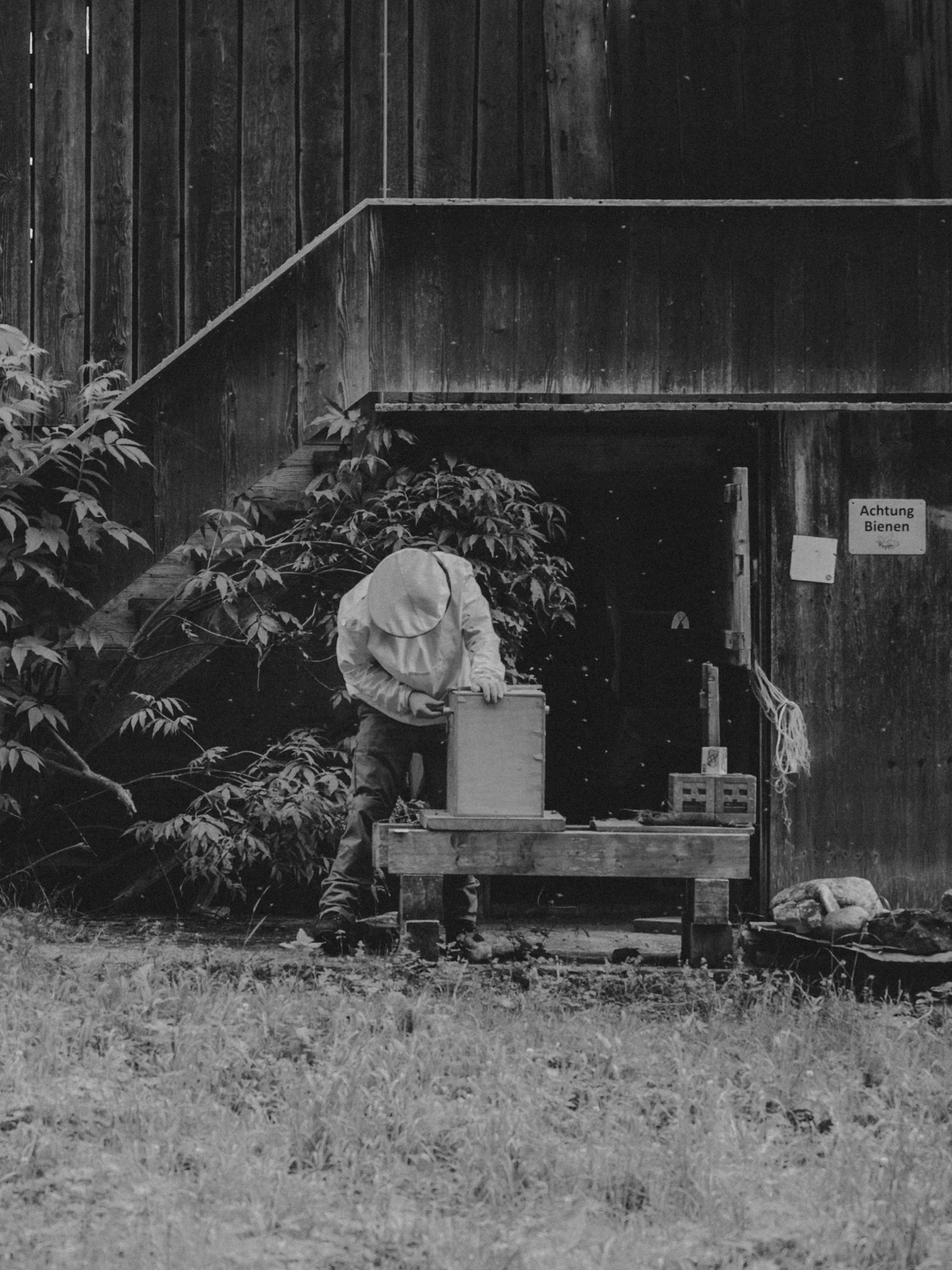 A person wearing a large hat working at a wooden table outdoors, surrounded by greenery and plants, with a wooden building in the background and a sign that reads 'Achtung Bienen'.