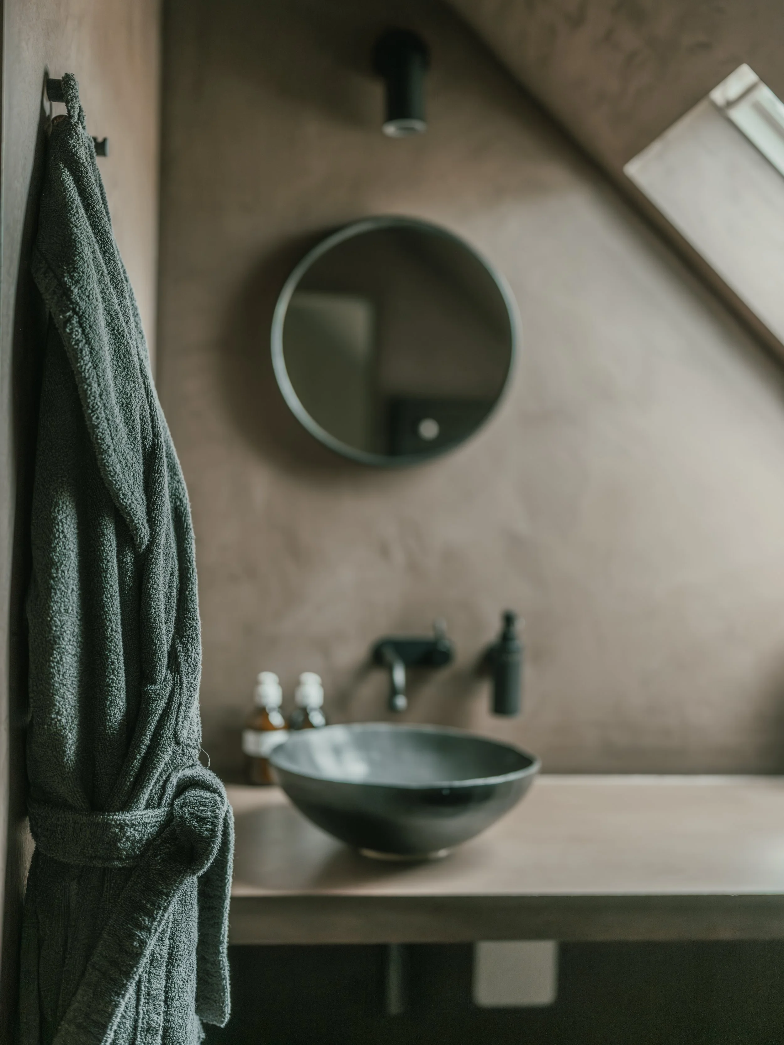 A bathroom with a gray towel hanging on a hook on the left side, a round mirror above a black vessel sink, a faucet and soap dispensers on the countertop, and a small window on the ceiling to the right.
