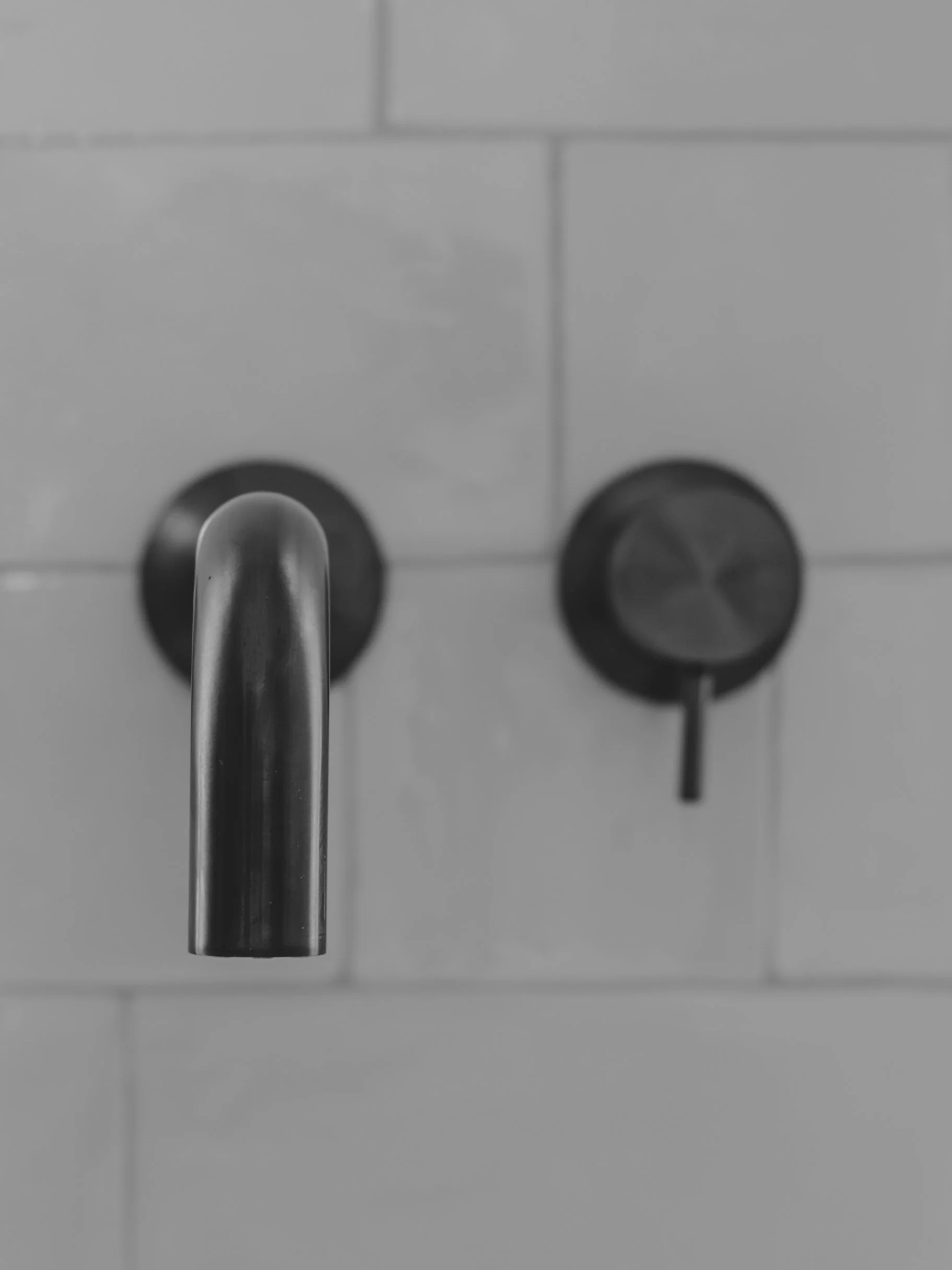 Close-up of a stainless steel faucet with a downward curved spout in front of a white tiled wall with black circular fixtures.