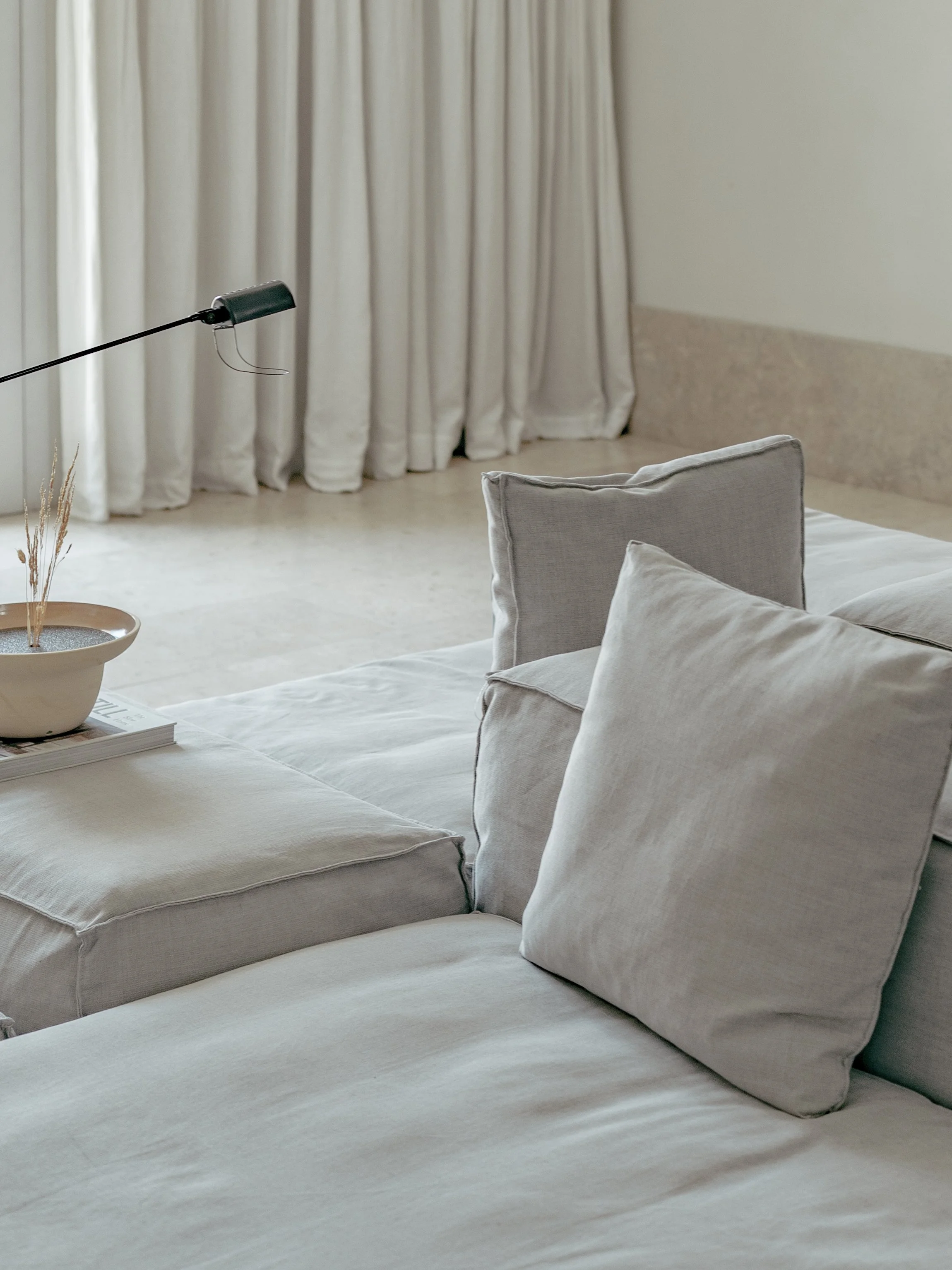A bedroom with beige curtains, a beige bed with a white bedspread, and beige pillows. There is a side table with a white book, a decorative bowl with dried plants, and a black reading lamp attached to the wall.