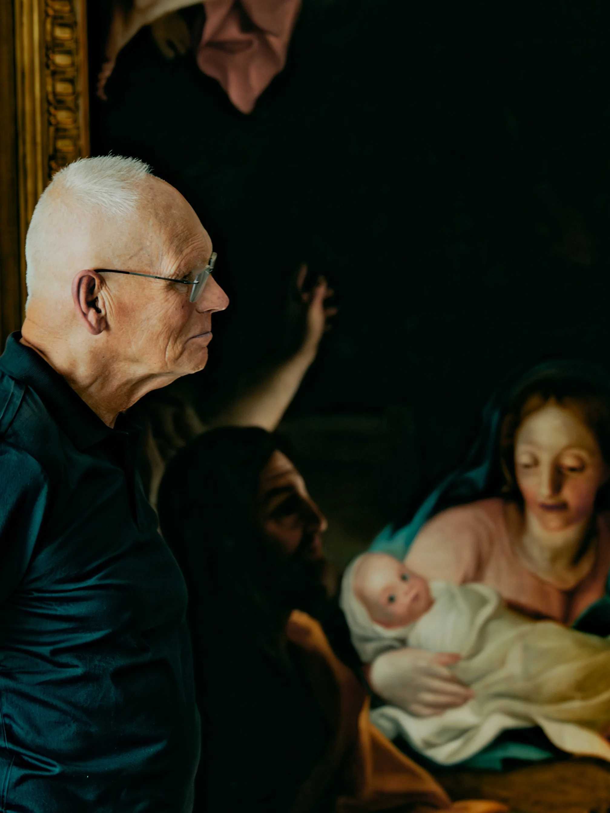 An elderly man with white hair and glasses is sitting, looking down, in front of a woman who is breastfeeding a baby. The scene appears to be in a dimly lit room, with other people visible in the background.