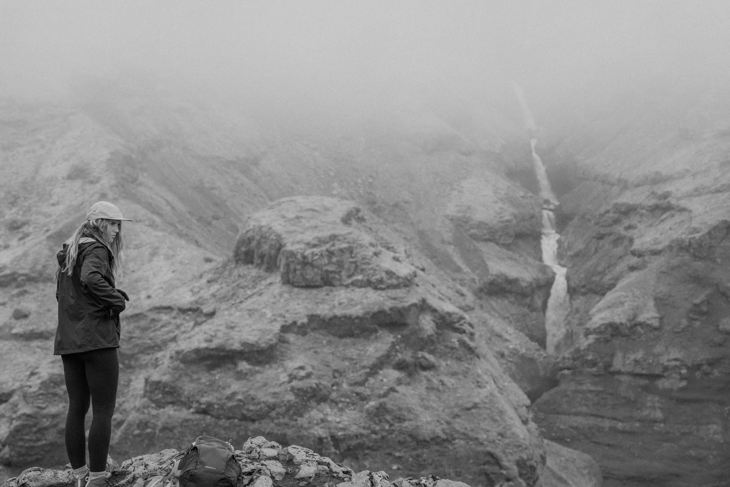 A woman in a jacket, leggings, and cap standing on rocky terrain in a mountainous environment with a waterfall in the background.