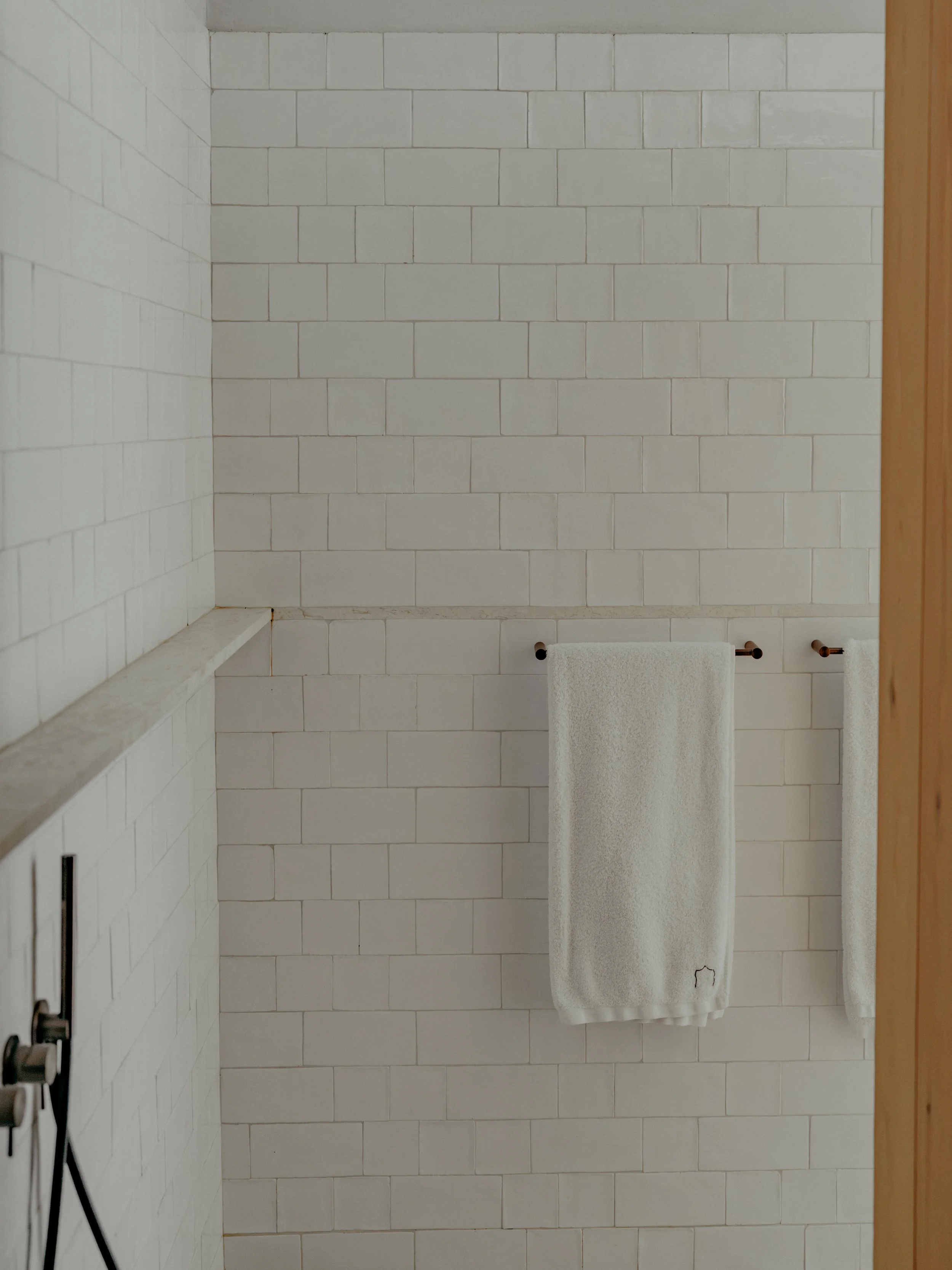 A bathroom with white tiled walls, two white towels hanging on a rack, and a partial view of a wooden door frame.