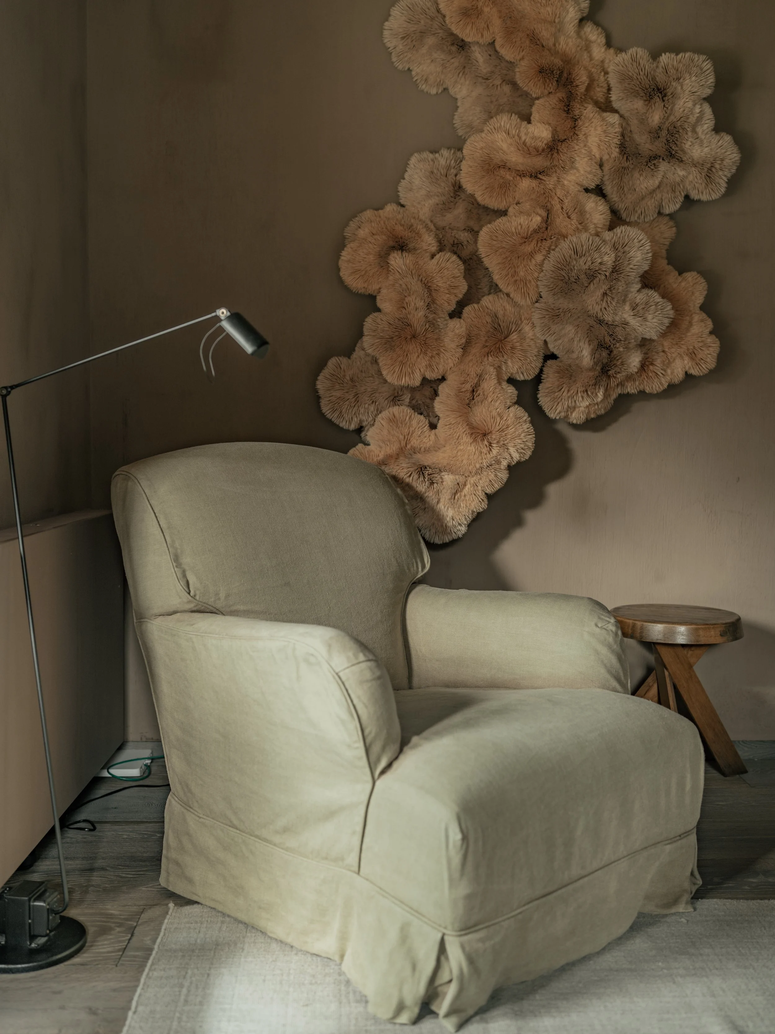 Cream-colored armchair with a floral wall art piece above it, next to a small wooden side table, situated in a cozy corner with wooden flooring and small rug.