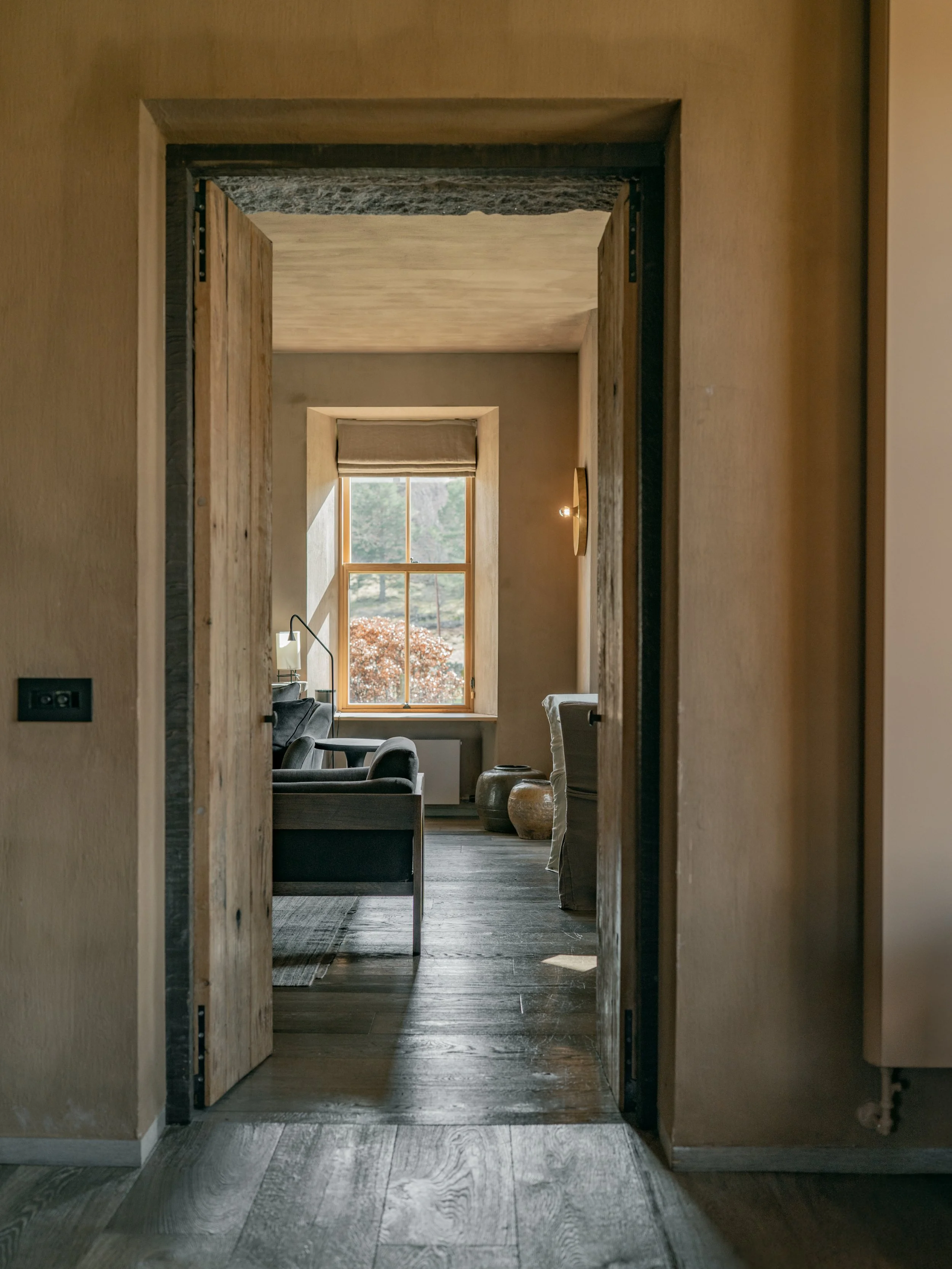 Interior view of a cozy room with a large window showing trees outside, furnished with comfortable armchairs, a side table, and decorative pottery, seen through a doorway with rustic wooden door frames.