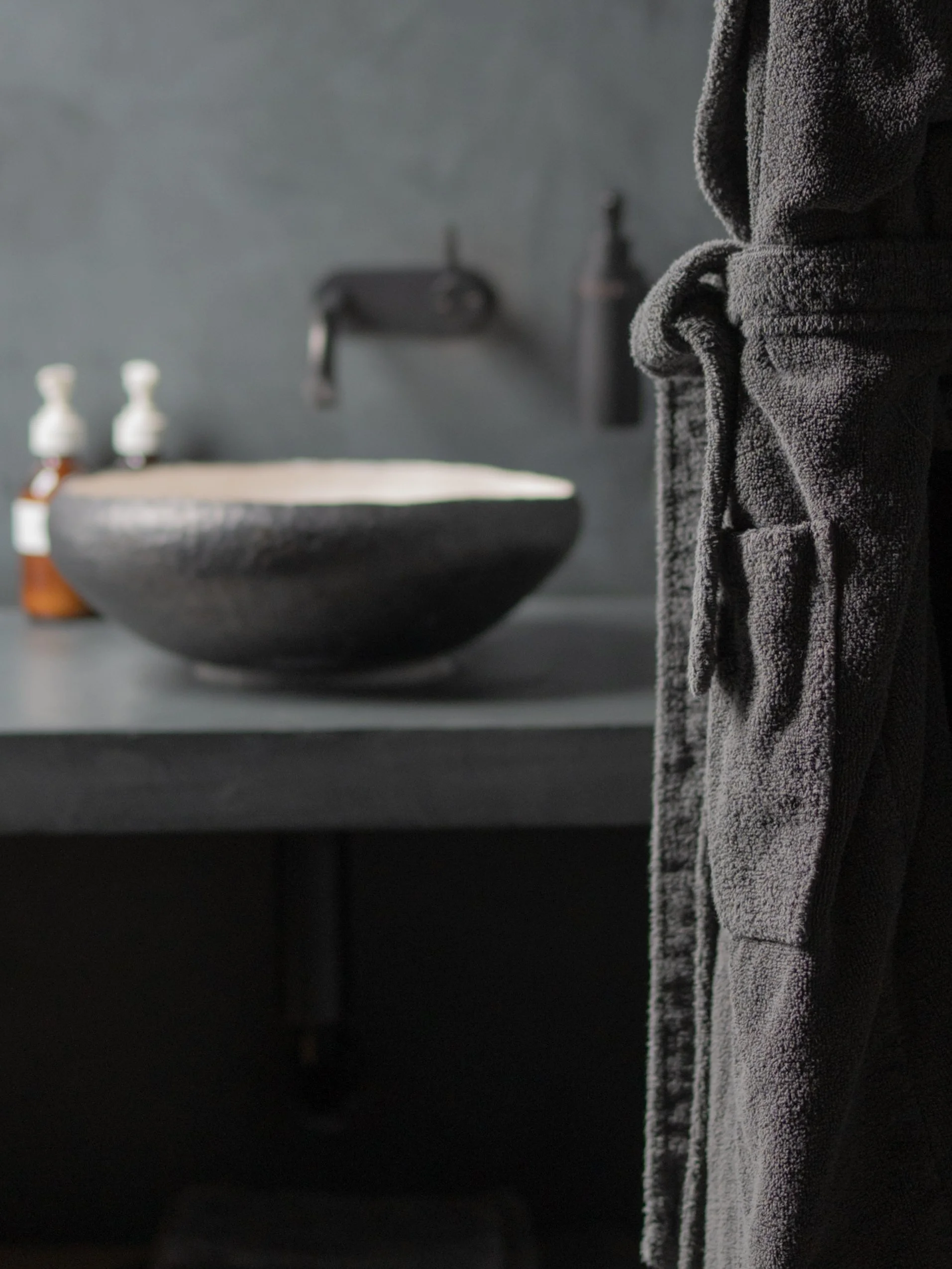Close-up of a dark gray towel hanging in a bathroom, with blurry background including a black sink and soap dispensers.
