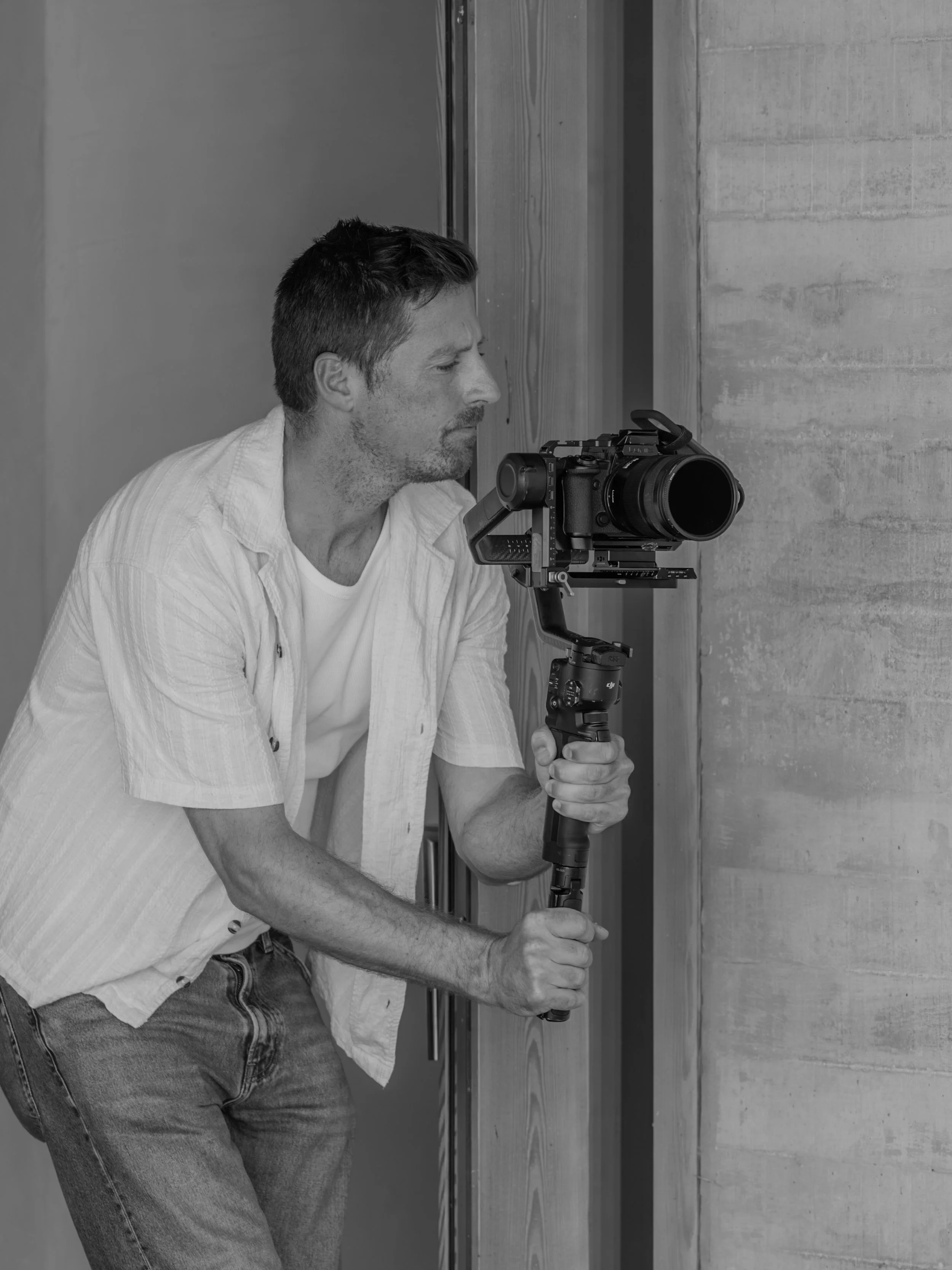 A man with short dark hair and a beard is operating a professional camera mounted on a stabilizer. He is wearing a light-colored shirt and jeans, and appears focused on filming or photographing. The background features a concrete wall and a reflectiv