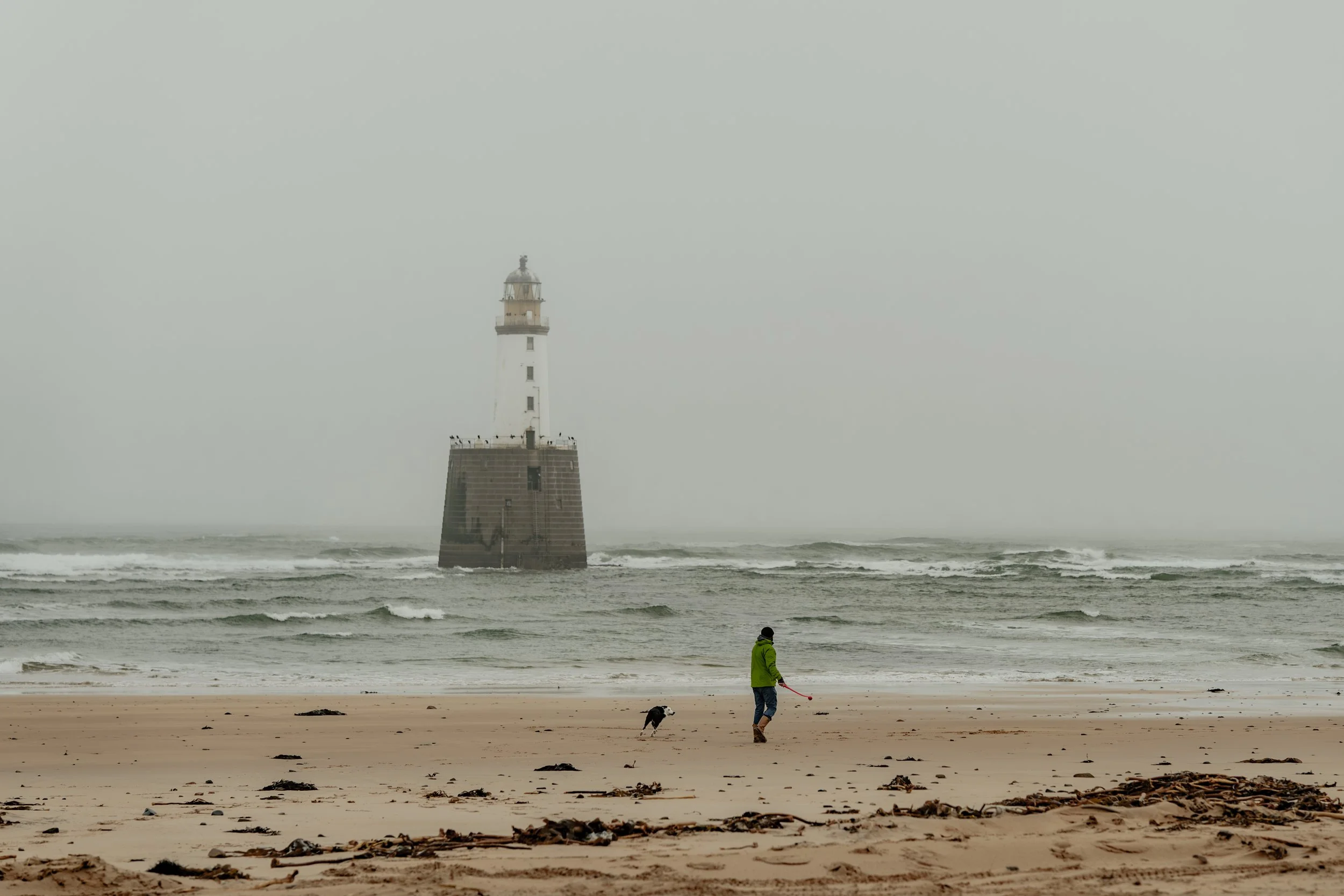 A person walking a dog along a sandy beach with a lighthouse in the background on a foggy day.