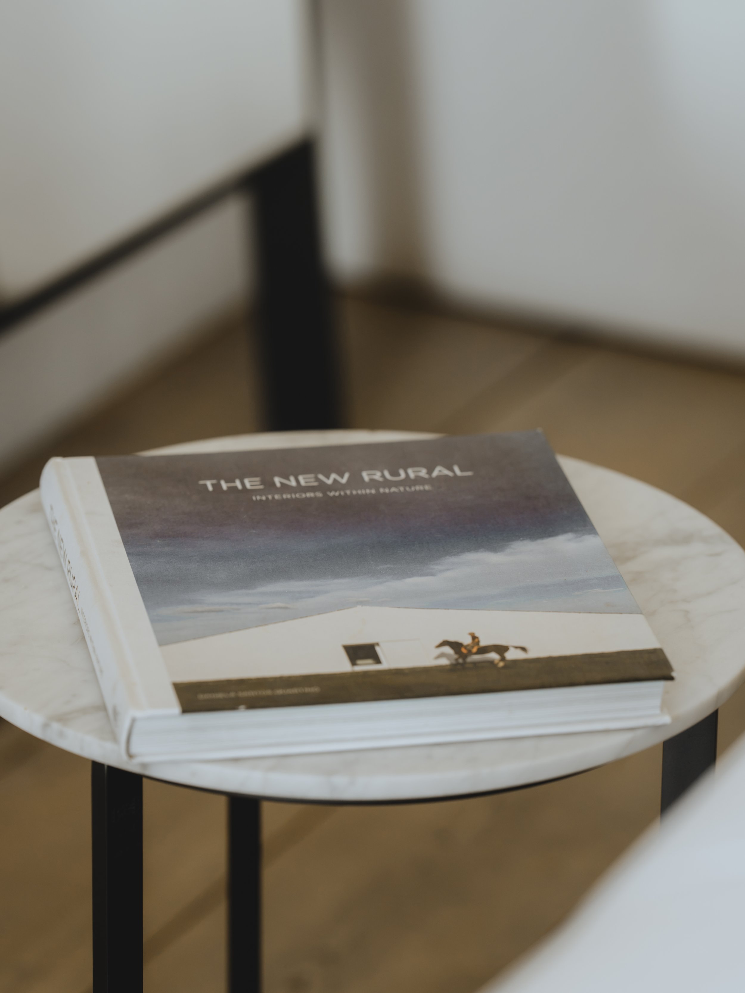 A book titled 'The New Rural' resting on a white table with a black metal leg, situated against a plain, white wall.