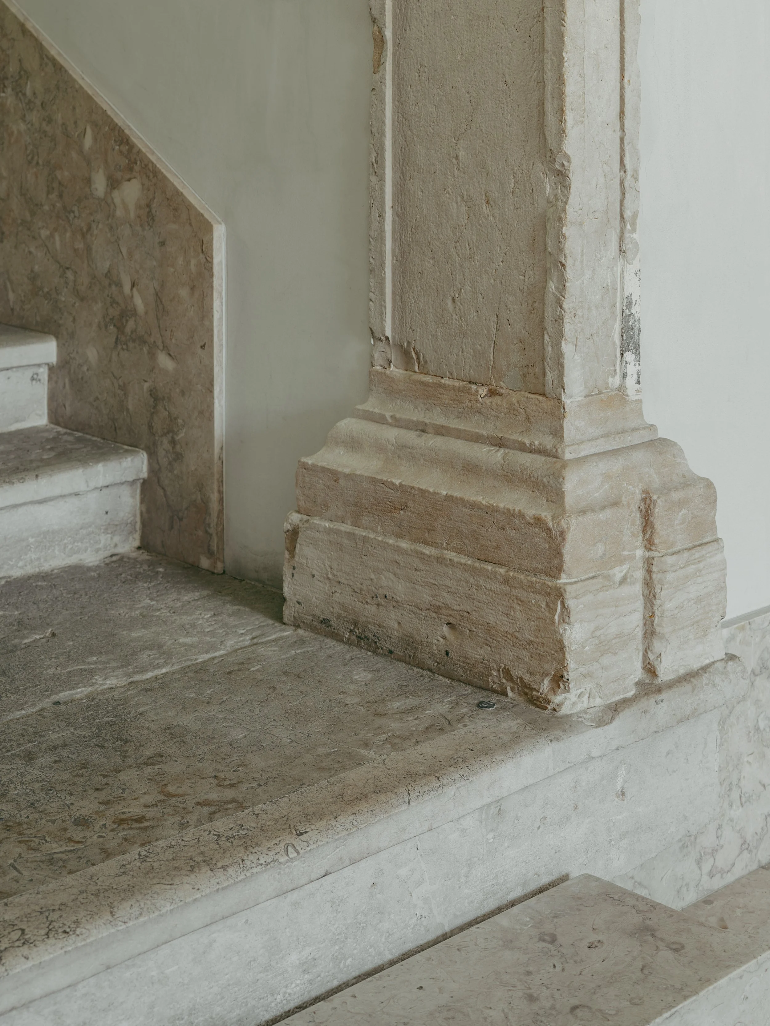 Close-up of a section of a wall with visible damage and broken plaster, revealing underlying stone and brickwork, next to a staircase made of beige stone or marble.
