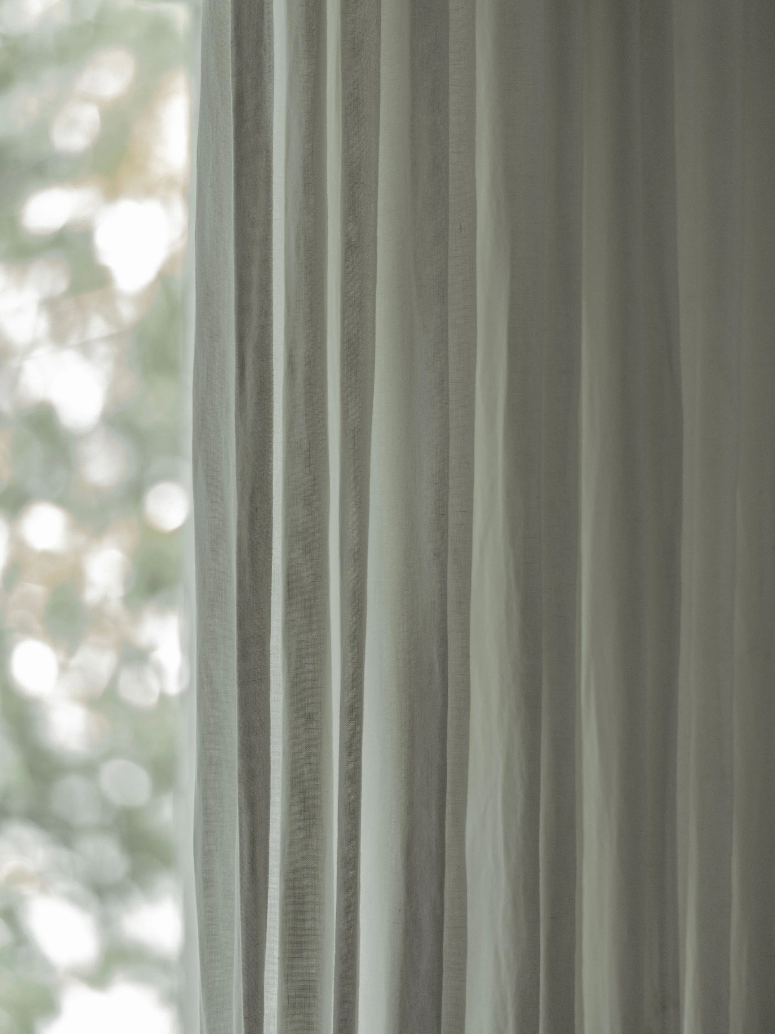 Close-up of sheer white curtains hanging in front of a window with daylight and blurred trees outside.