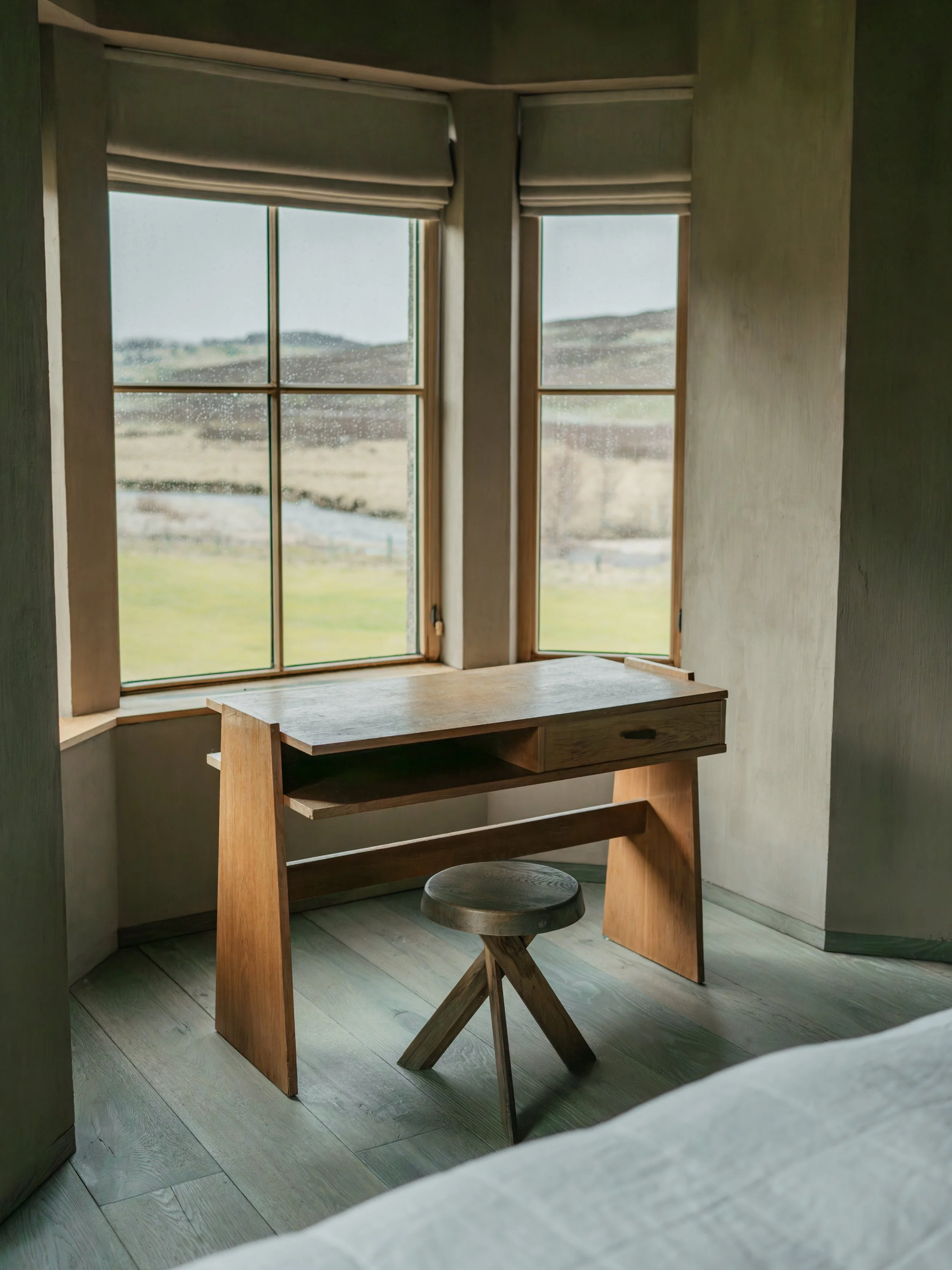 A wooden desk and stool near two windows with closed shades, overlooking a landscape of hills and fields.