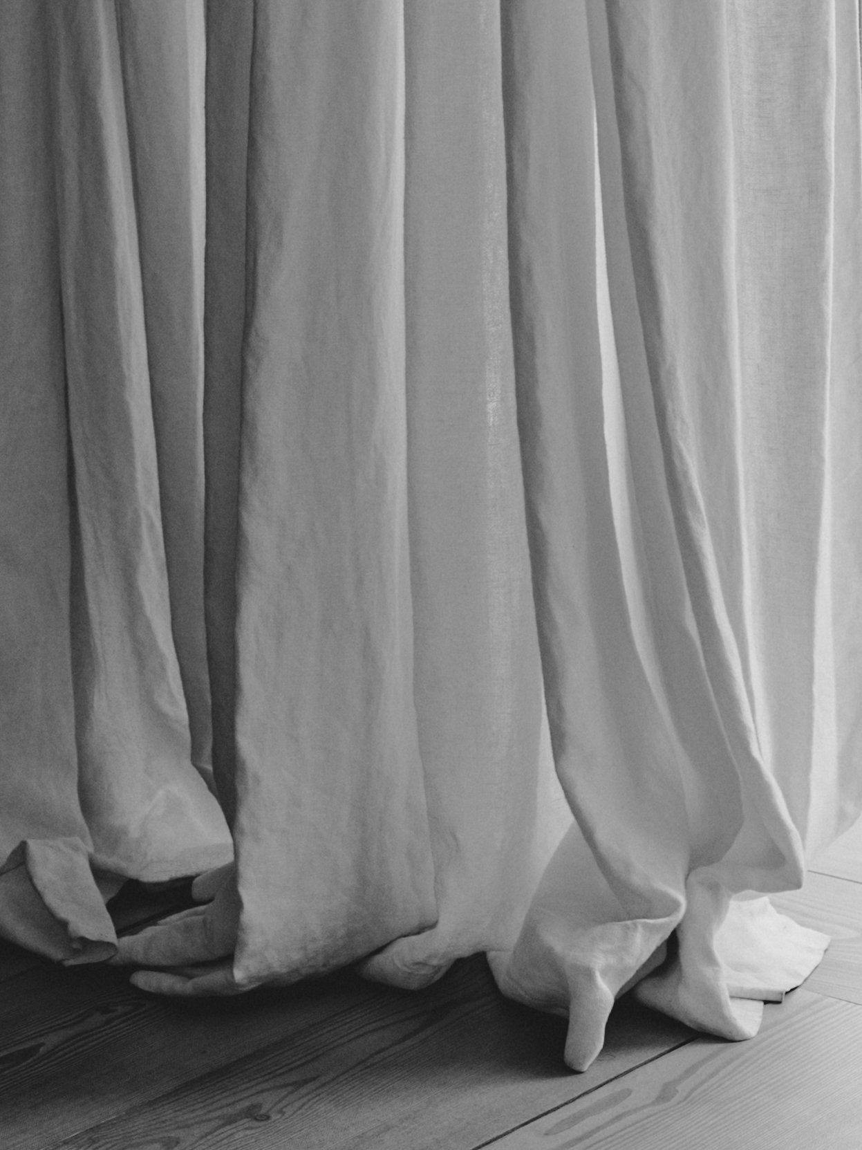 A white curtain hanging from a window or rod, pooling slightly at the bottom on a wooden floor.