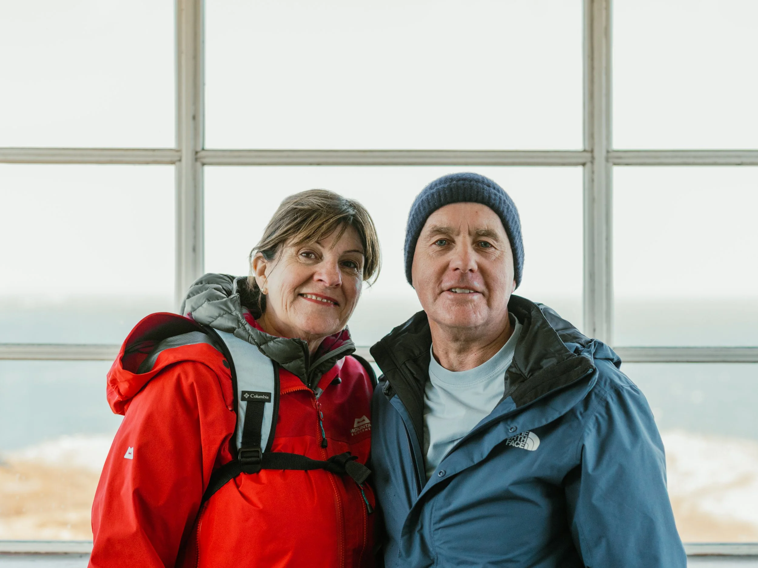 Two middle-aged adults, a woman and a man, standing close together inside a building with large windows behind them. The woman has short brown hair, is smiling, and is wearing a red jacket with a backpack. The man has short gray hair, is wearing a gr