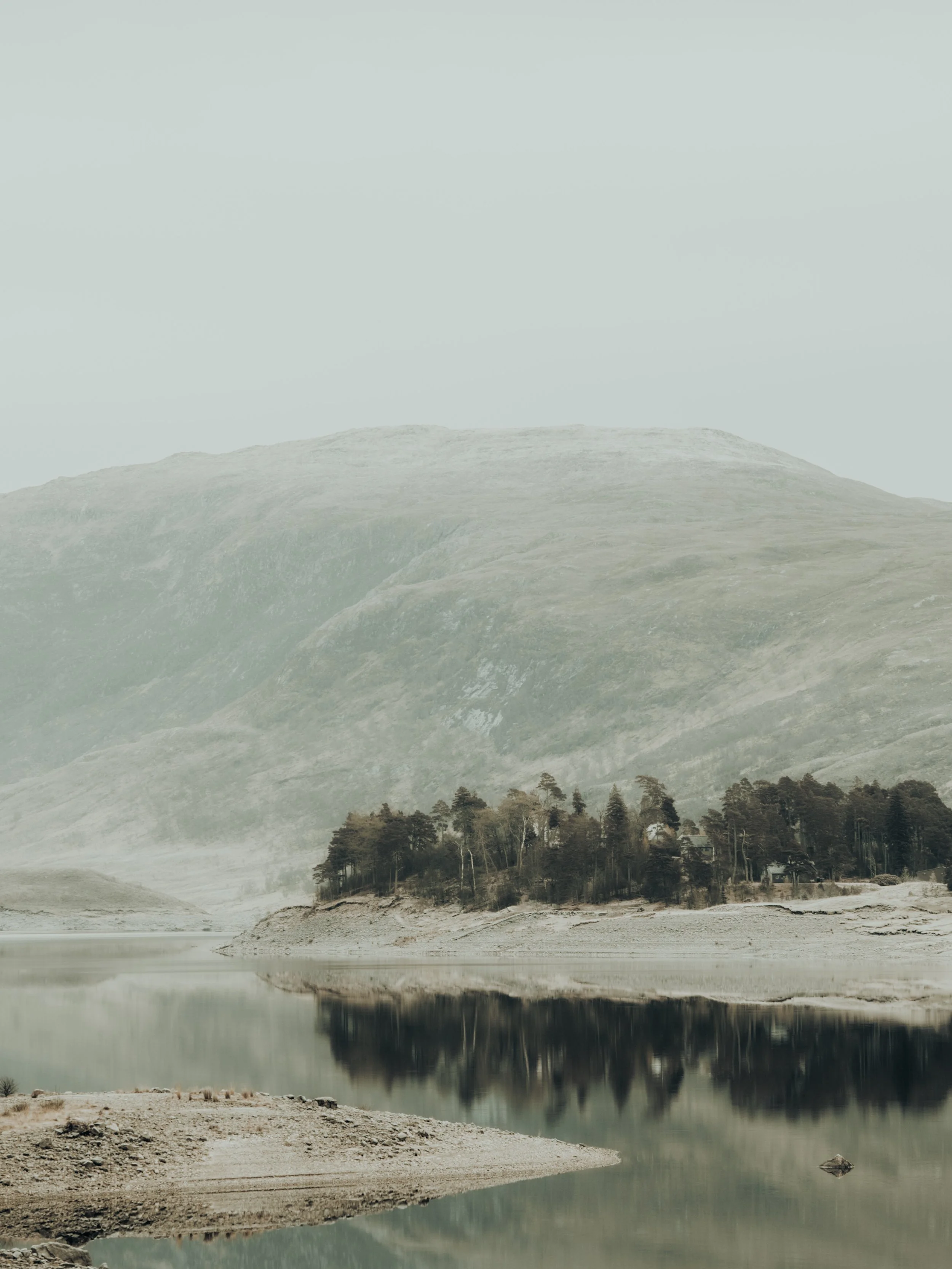 A moody morning at a loch in the Scottish highlands in Scotland with mountains, water, trees and mist