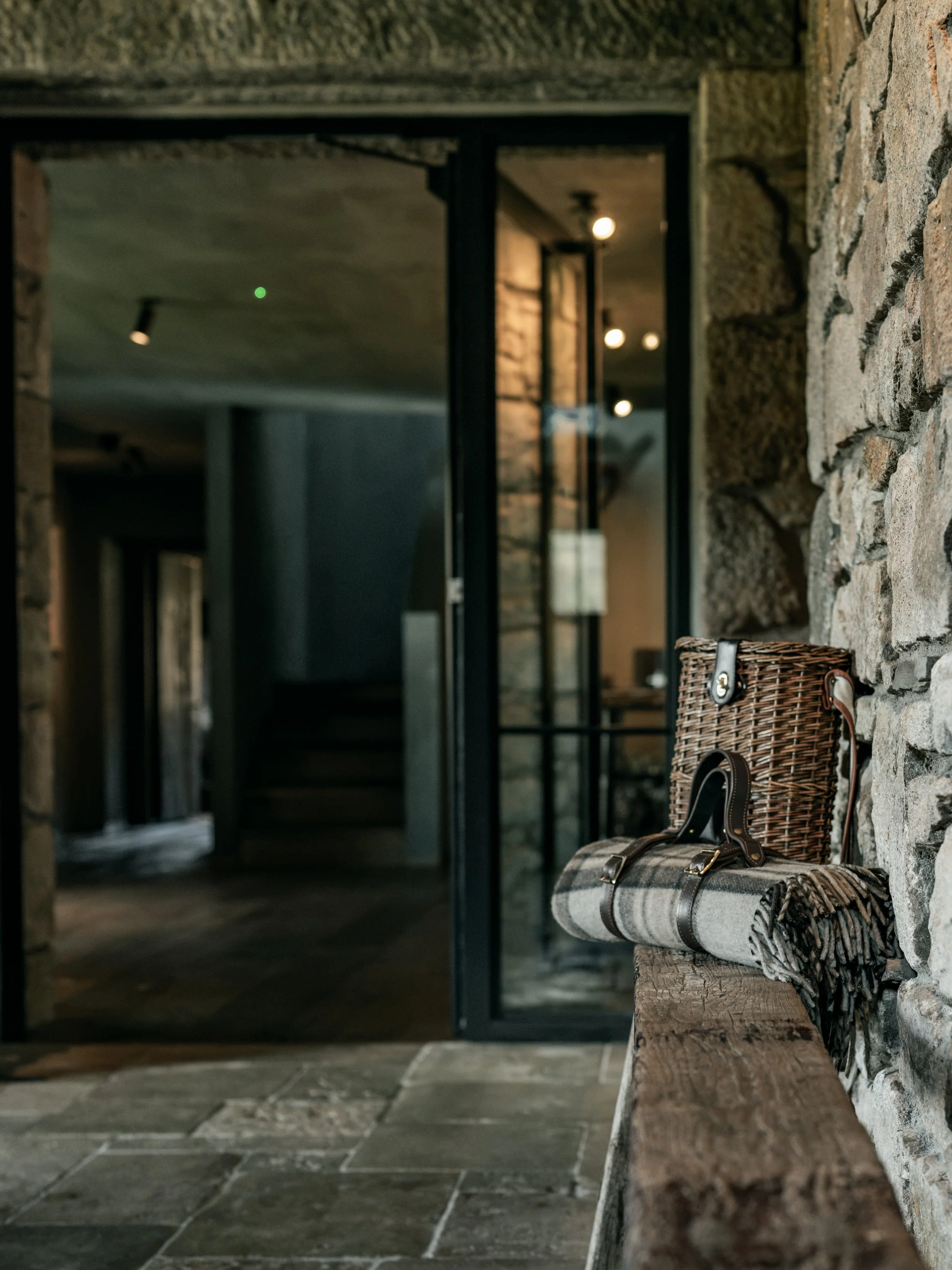A wooden bench against a stone wall with a woven purse, glasses, and a plaid blanket resting on it.