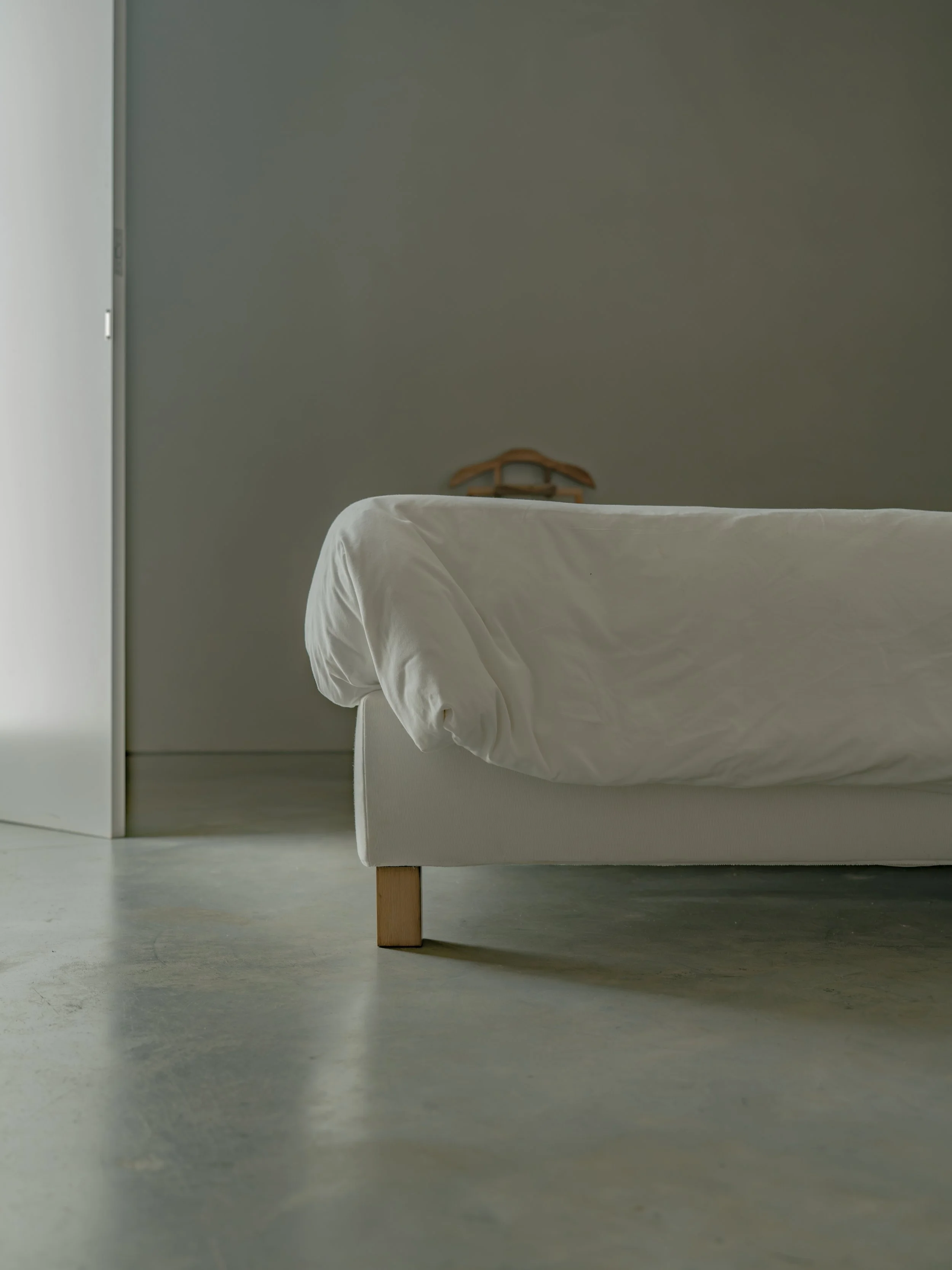Part of a bed with white sheets in a minimalist room with a green wall and a wooden hanger on the wall.