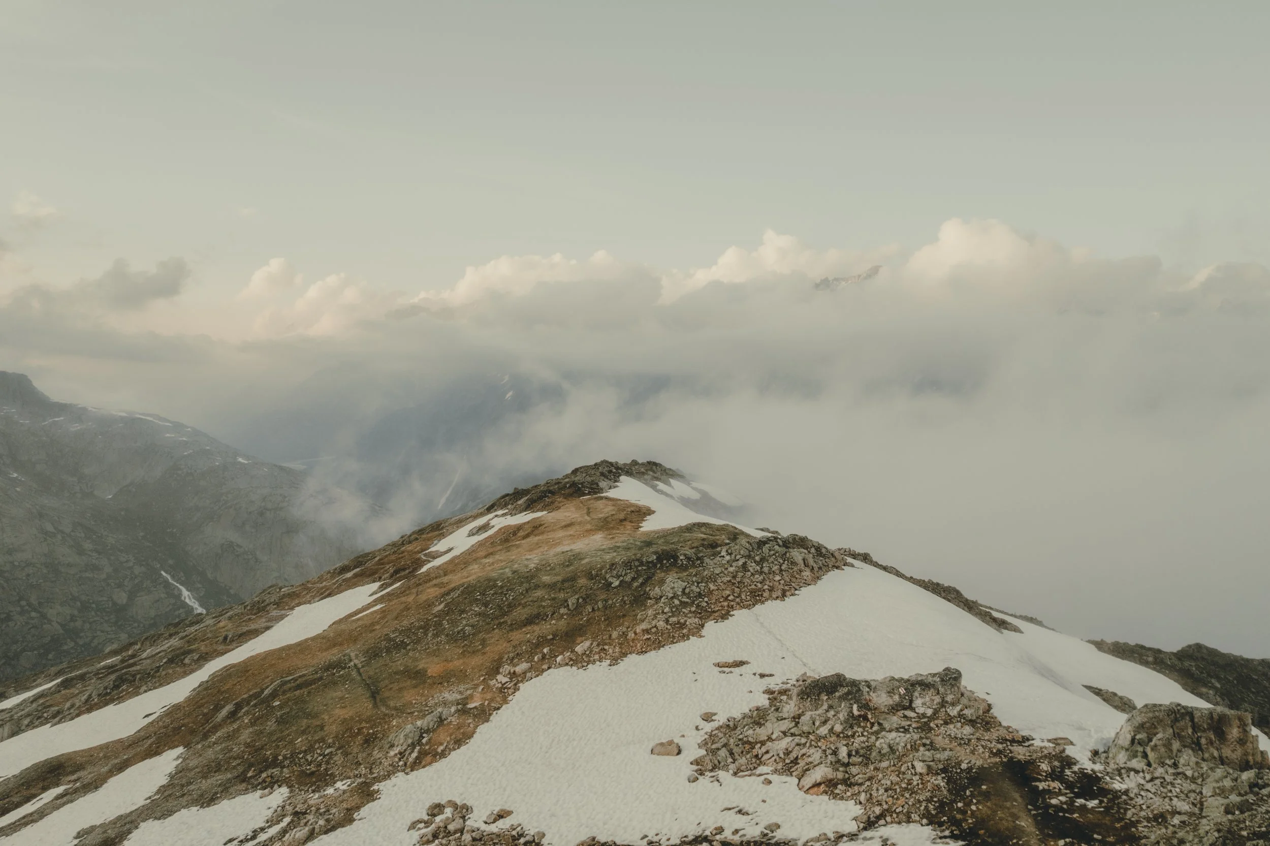 Mountain landscape with patches of snow on rocky terrain, surrounded by clouds and fog, with distant mountain peaks partially visible.