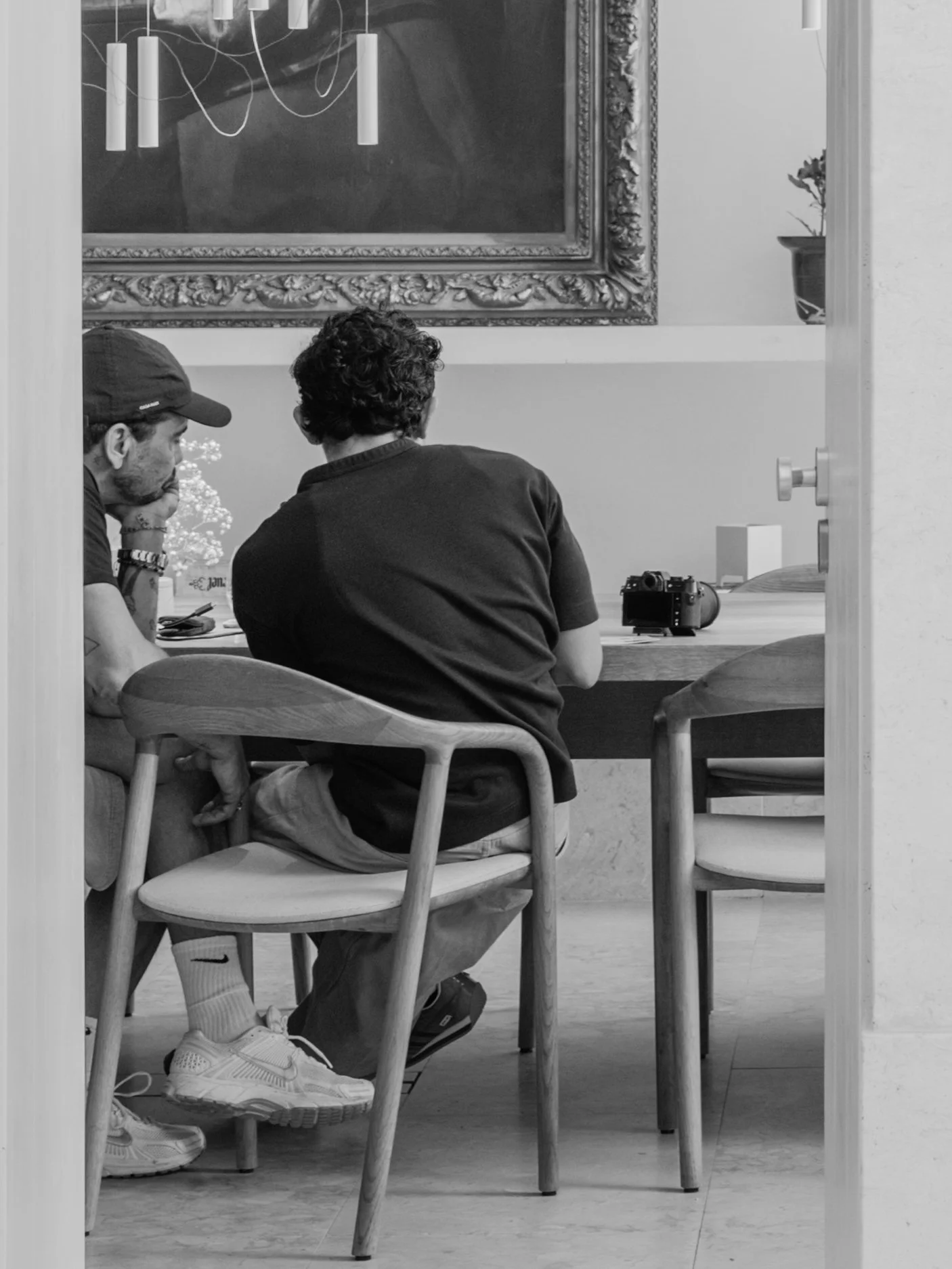 Two young men sitting at a wooden table, viewed through a doorway, with a large ornate framed mirror and a potted plant in the background.