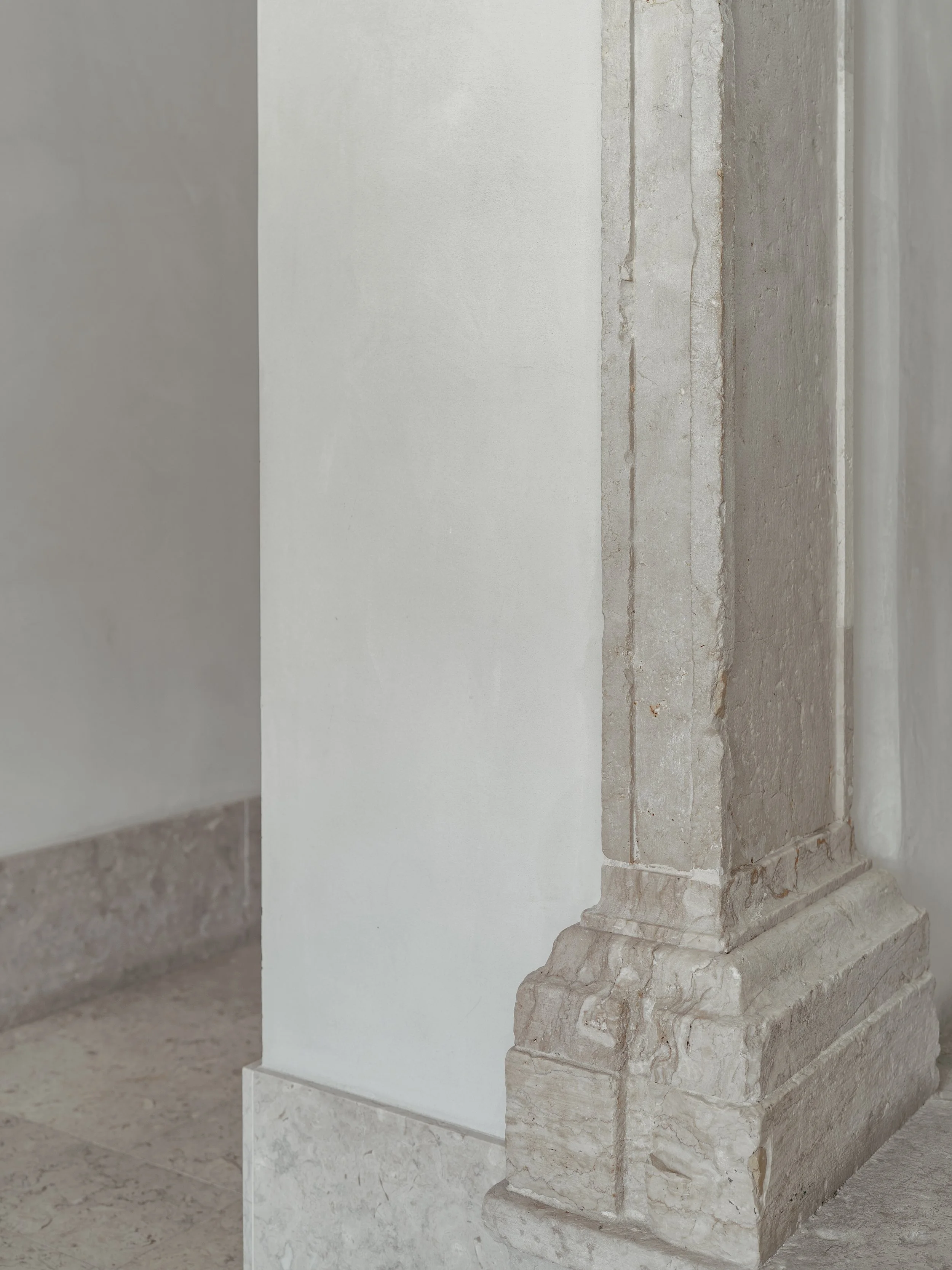 A decorative ancient stone sculpture of a standing figure, possibly a statue, carved from light-colored stone with intricate details, displayed indoors against a white wall.
