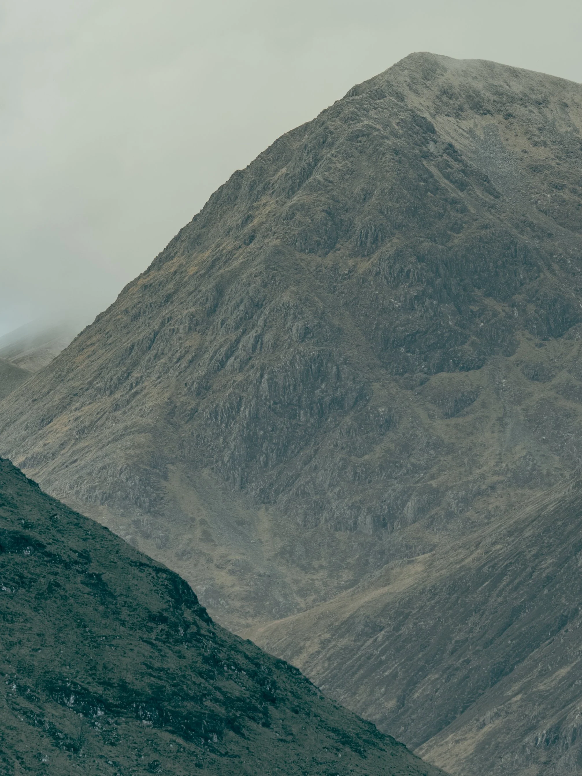 A mountainous landscape with rocky, rugged terrain and steep slopes, under a cloudy sky.