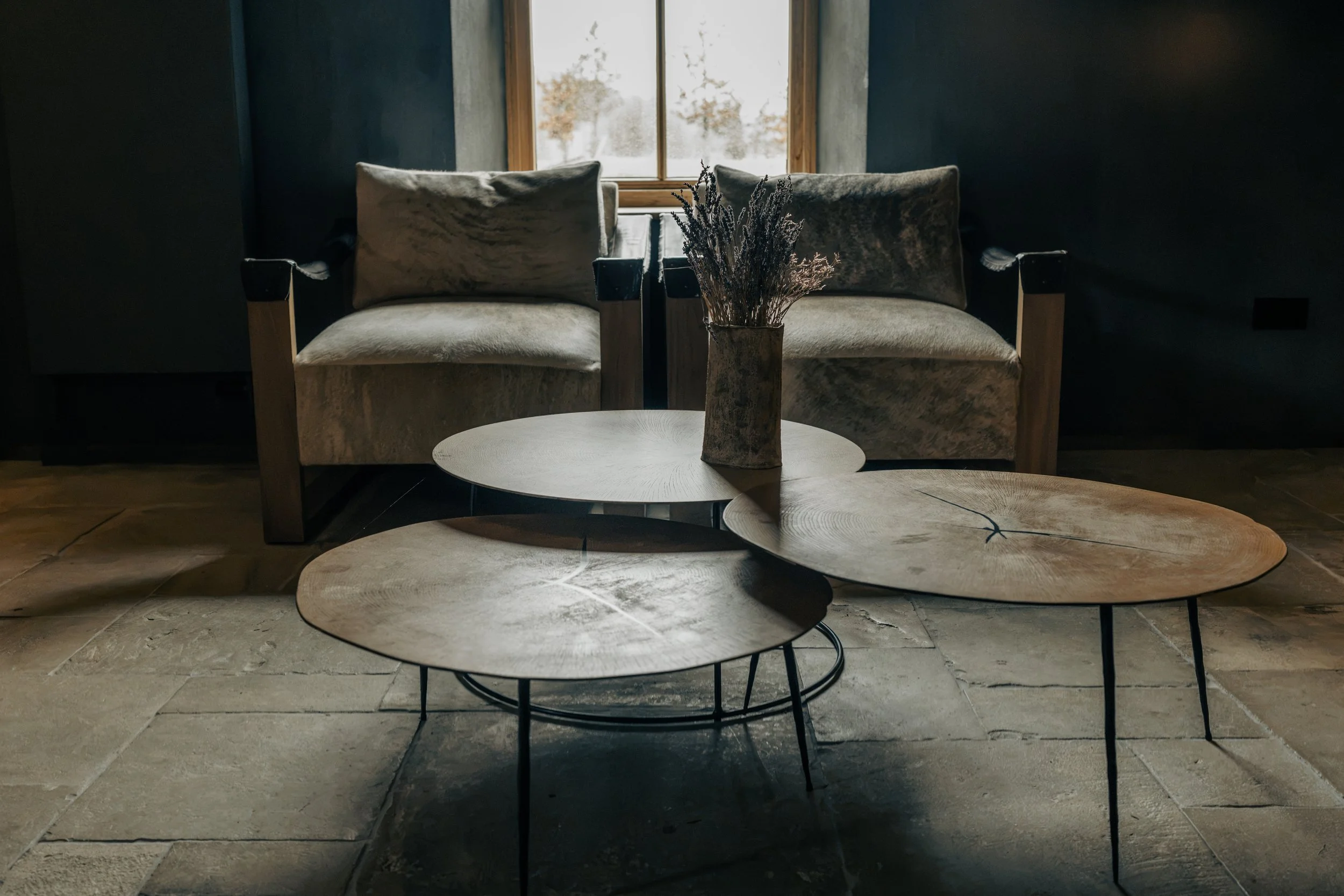 A cozy living room with a window showing outdoor trees, featuring a small sofa with pillows, a wooden vase with flowers, and round wooden coffee tables with metal legs on a stone floor.