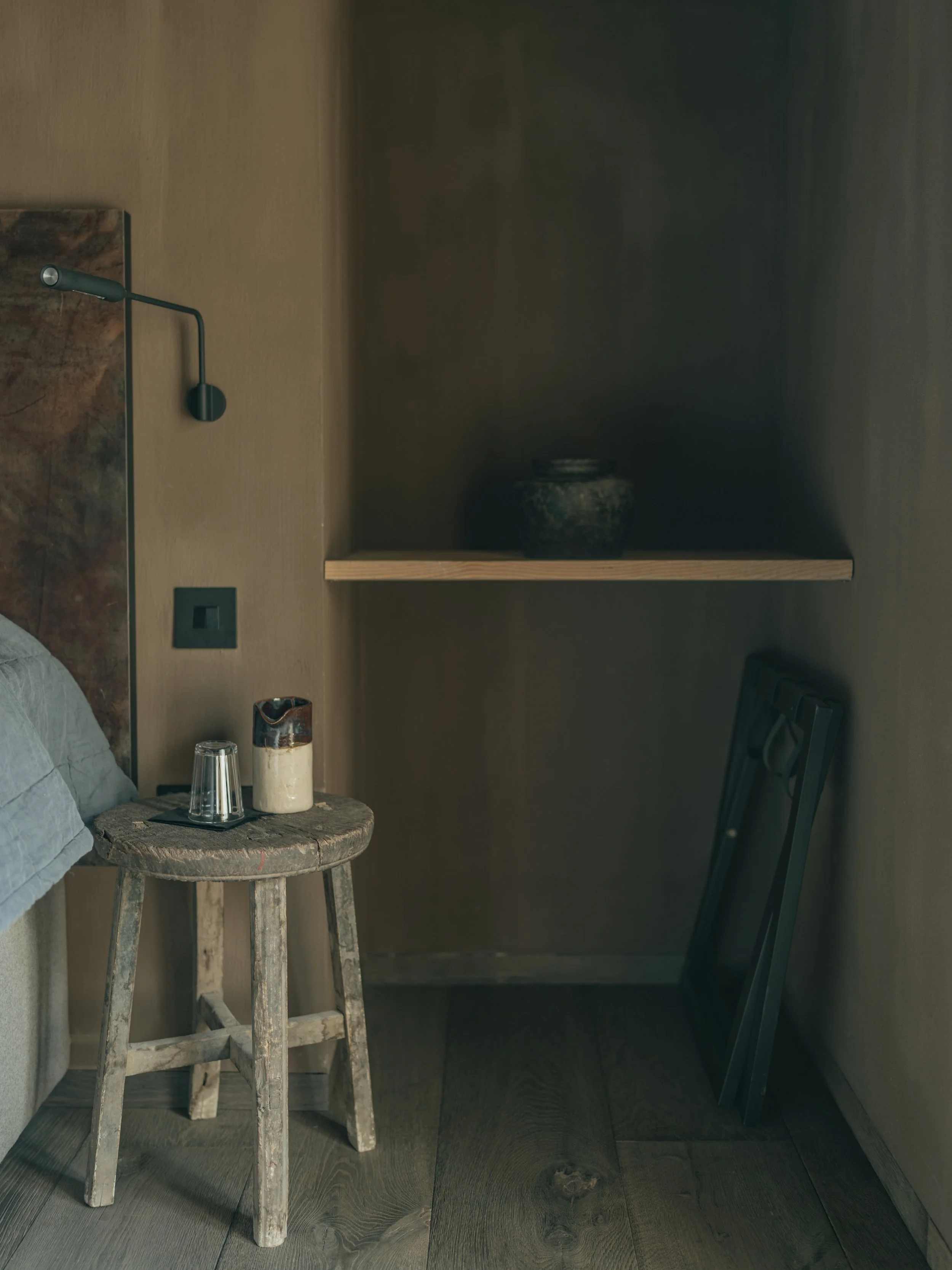 A rustic wooden stool with a glass, a ceramic cup, and a ceramic pitcher on top, positioned next to a bed with linen. To the right, there's a black chair leaning against a wall, and behind the stool, a wall lamp and electrical outlet are visible. A d