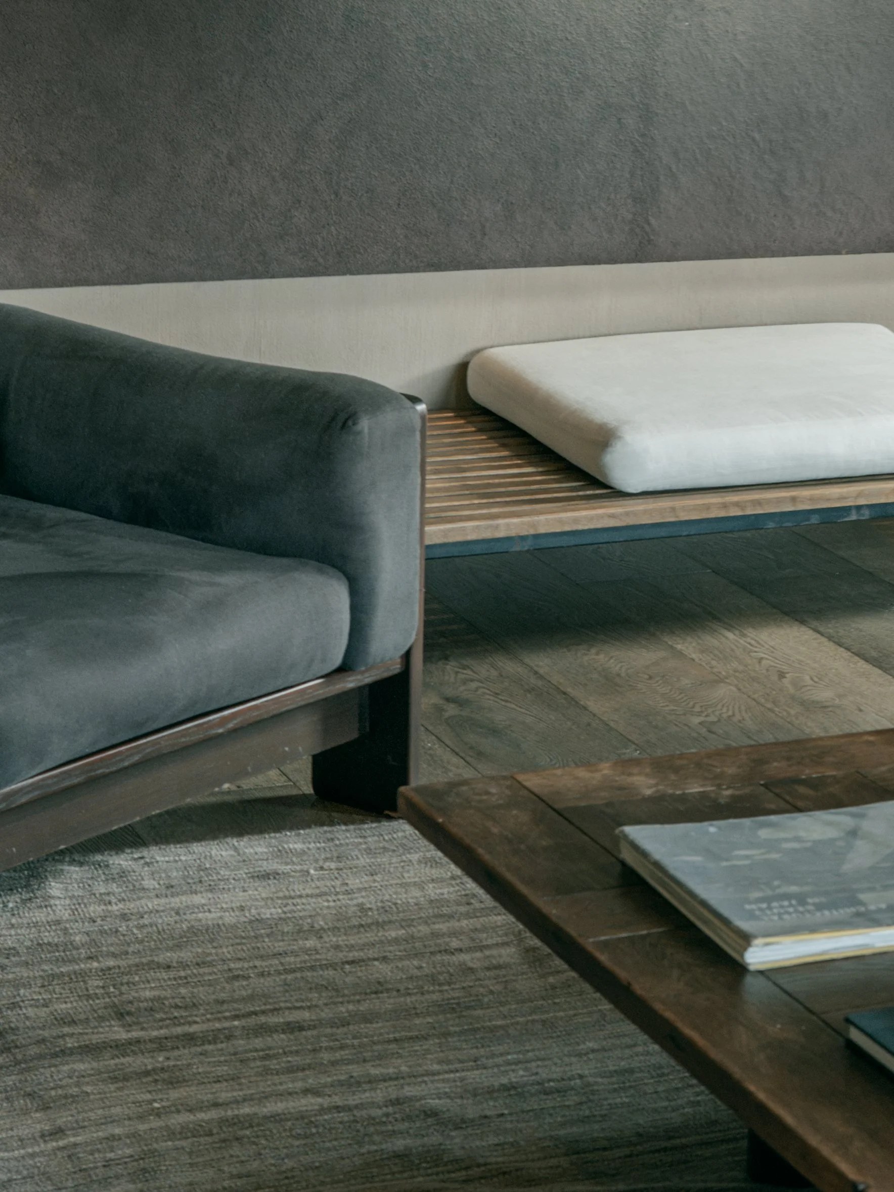Part of a living room with a dark gray sofa, a wooden coffee table with a magazine or book, a light-colored bed or cushioned bench, and a textured gray wall.