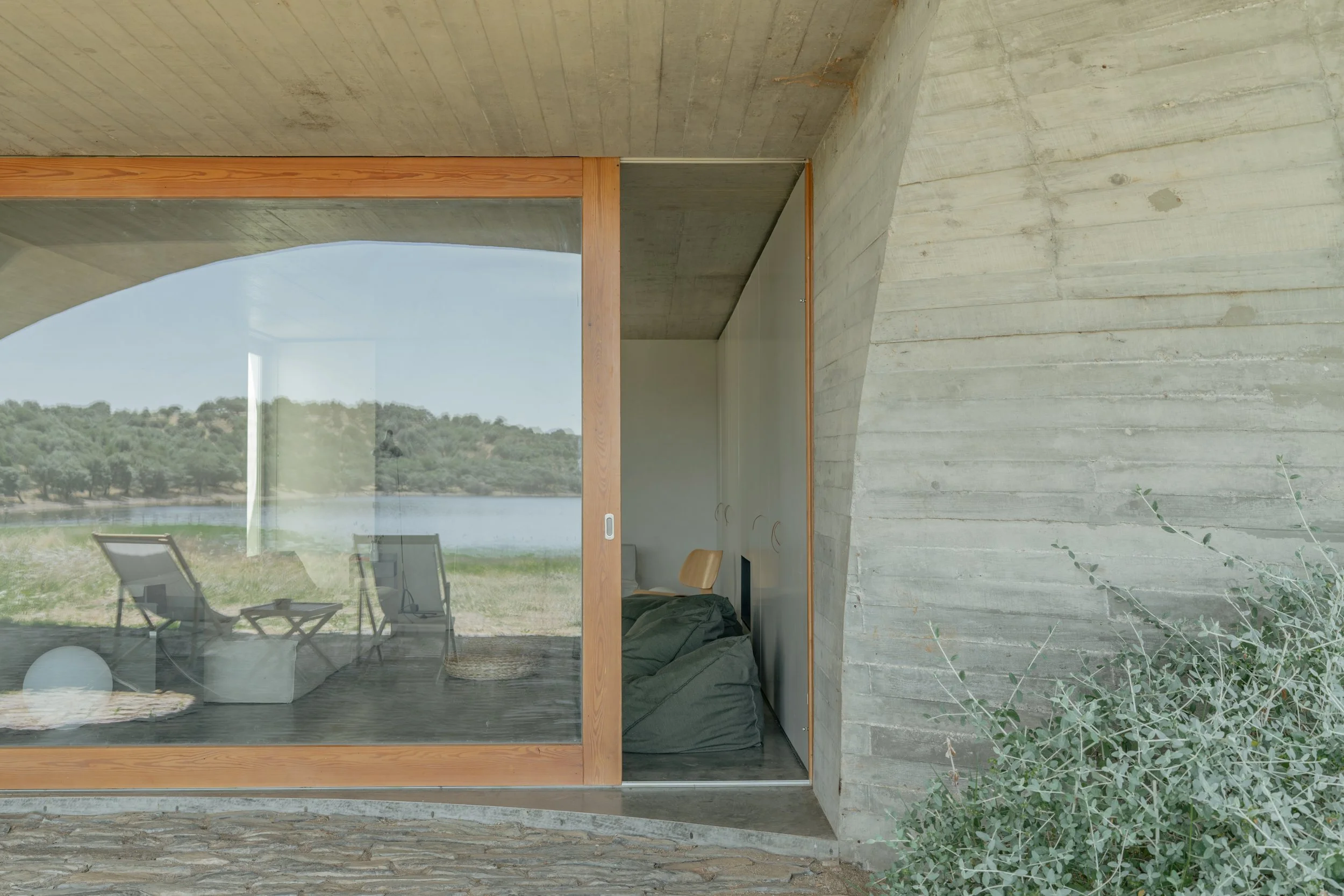 Interior view of a modern building with concrete walls, featuring large glass windows showing outdoor landscape. The room includes chairs and a bed with bedding, and has a wooden and concrete architectural design.