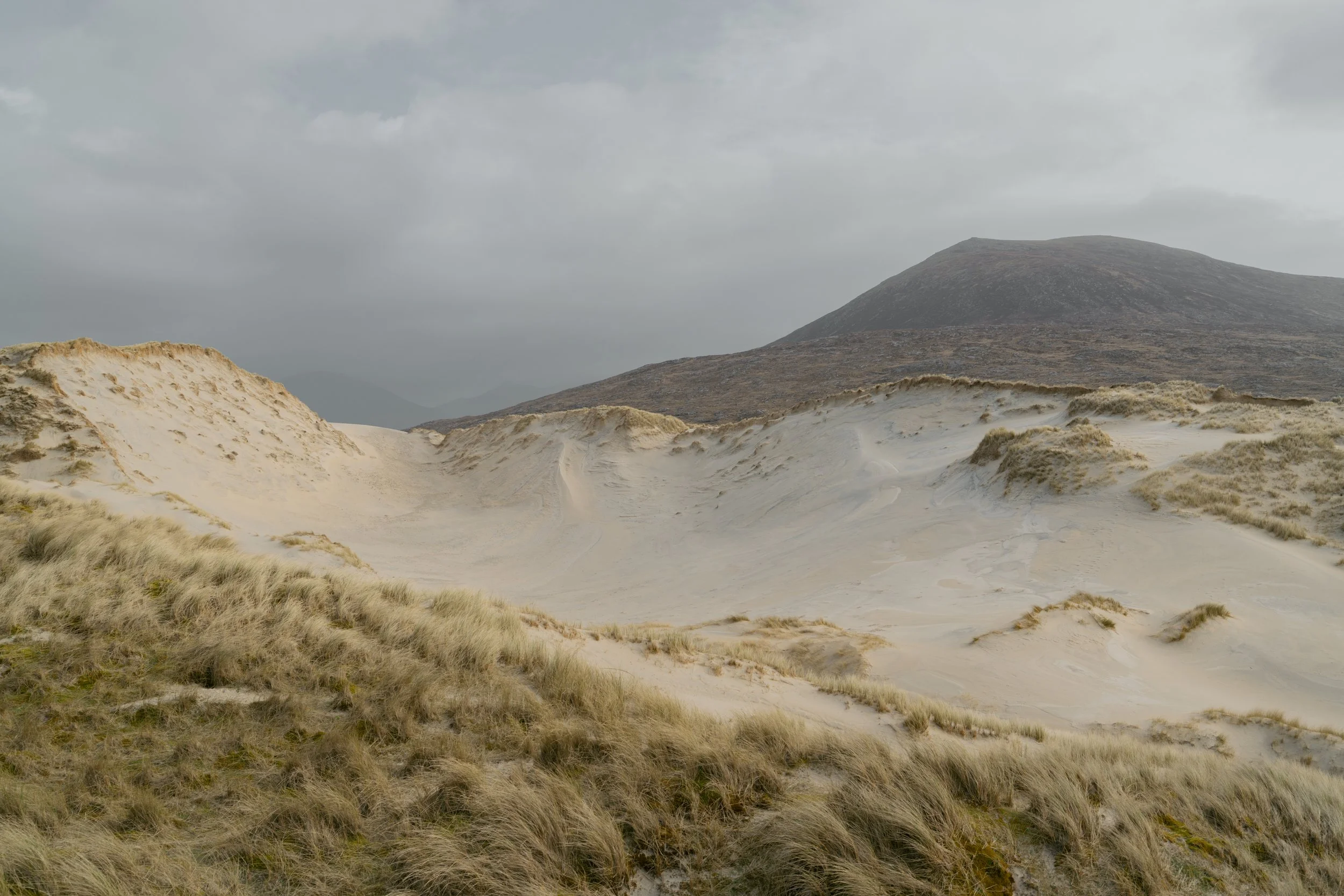 A sandy landscape with dunes in the foreground and a mountain in the background under a cloudy sky.