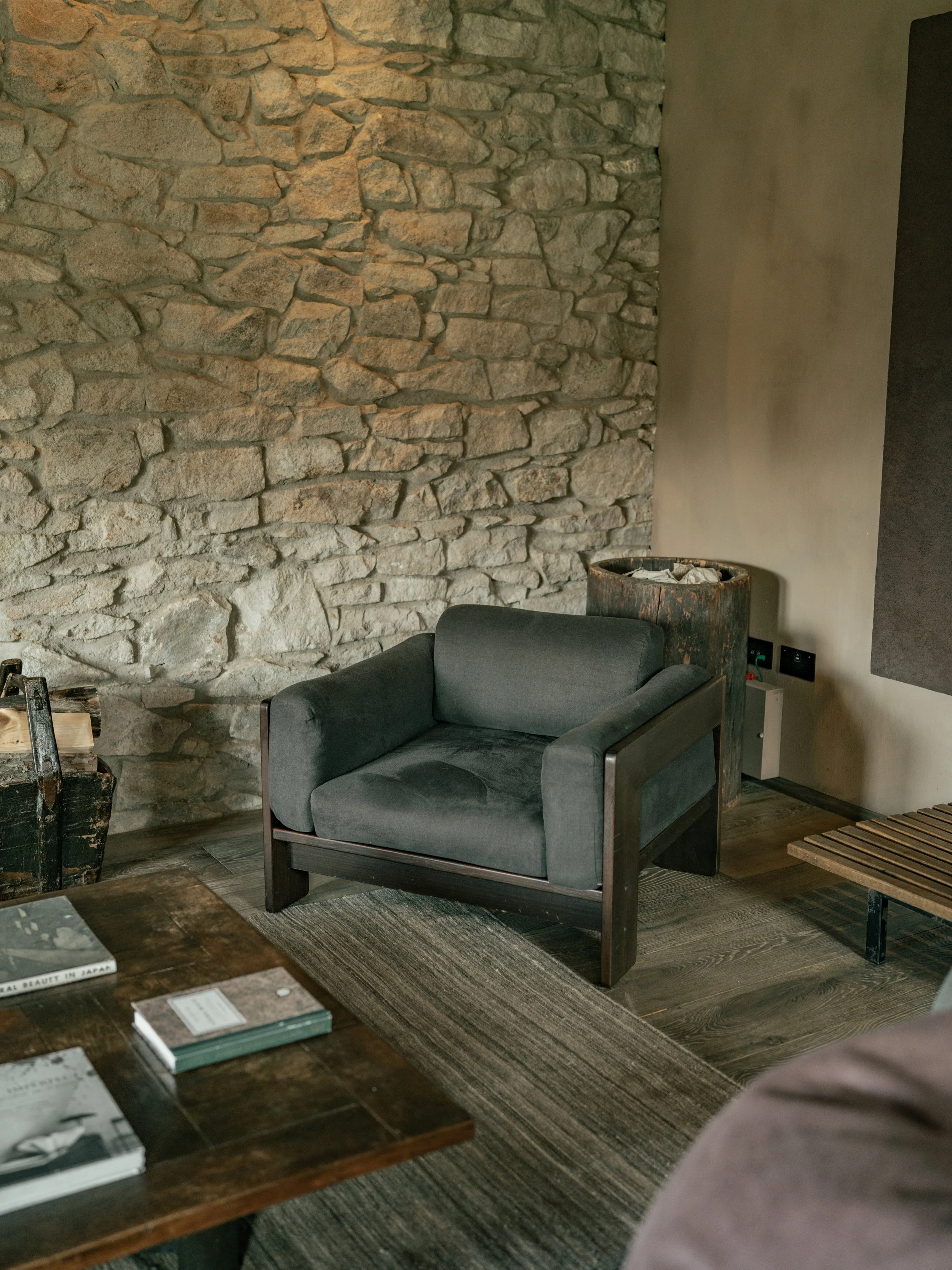 A seating area with a dark gray armchair positioned in front of a beige exposed brick wall, with a rustic barrel and a piece of furniture nearby, and a wooden table with books and magazines in the foreground.