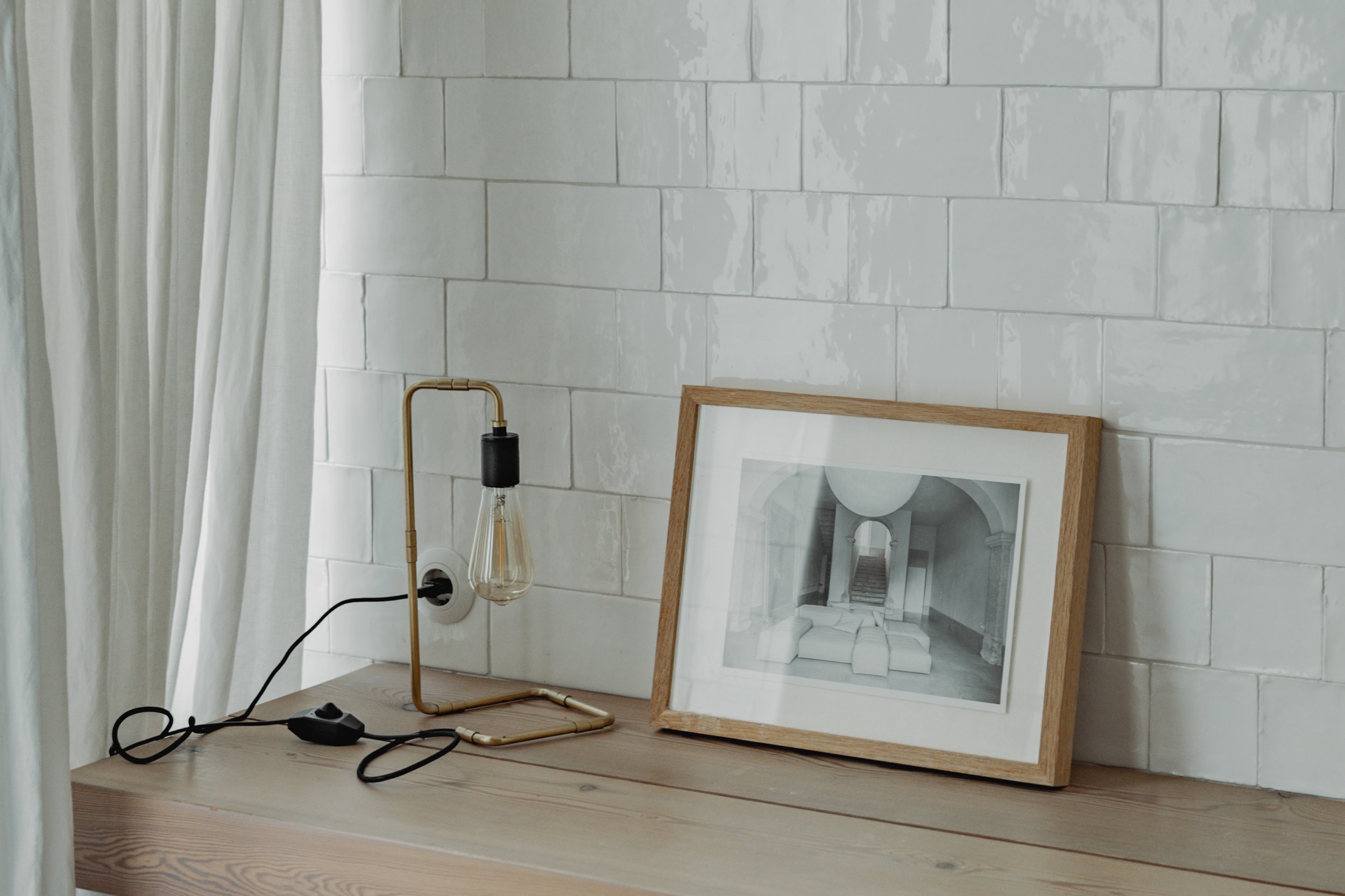 A corner of a room with white tiled wall and light wood surface. There is a framed black-and-white photo leaning against the wall, a modern table lamp with a black socket and brass stand, and a white curtain hanging to the side.