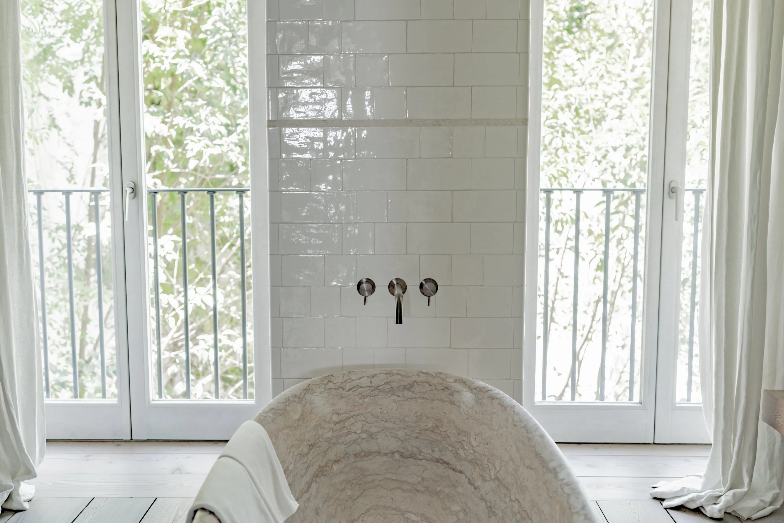 View of a bathroom with large windows and a white tiled wall, a bathtub in the foreground, and a balcony with a railing outside the window.
