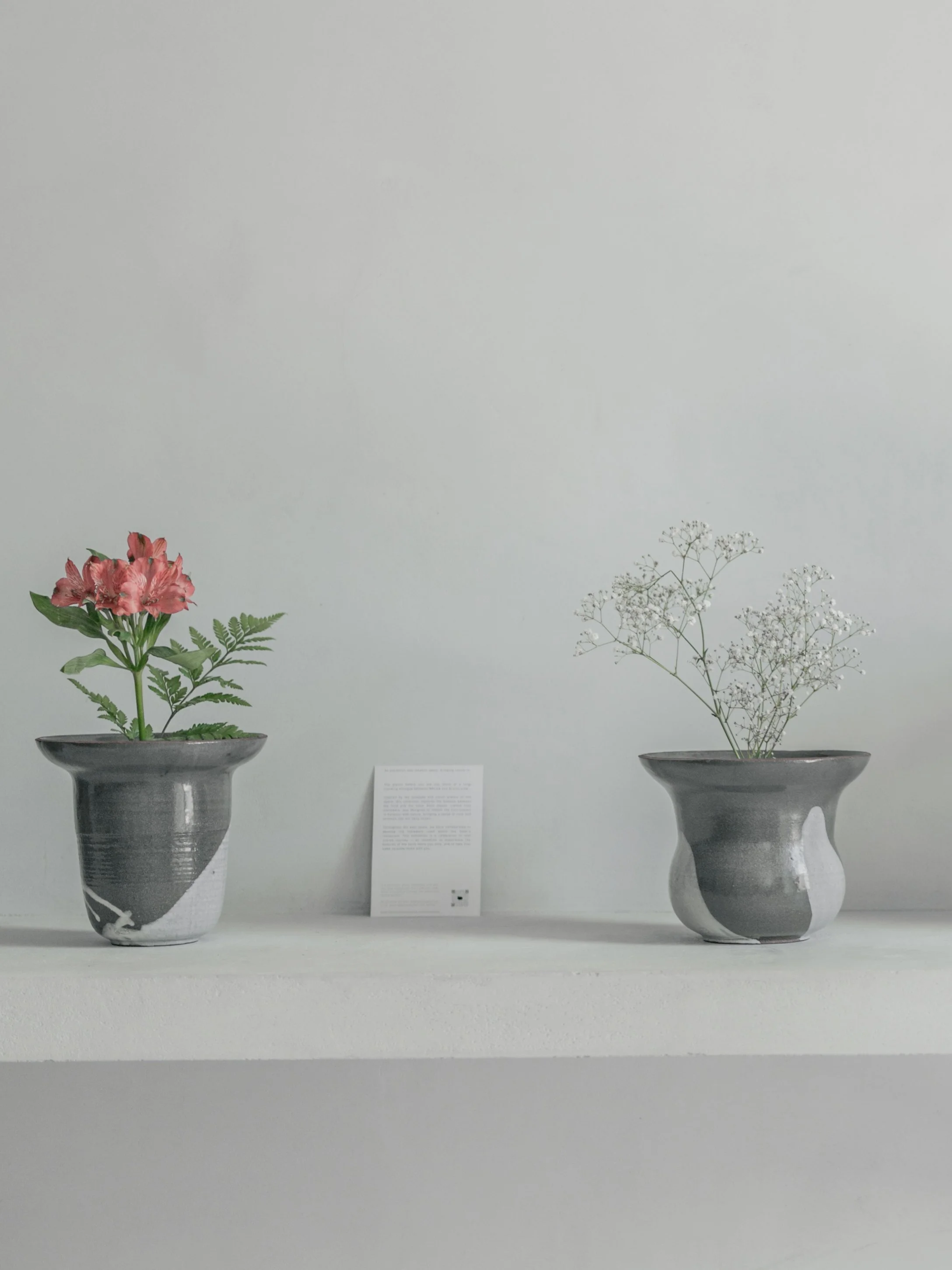 Two vases with floral arrangements on a white surface against a light gray wall. The left vase contains pink flowers and green foliage, the right vase contains delicate white baby's breath flowers.