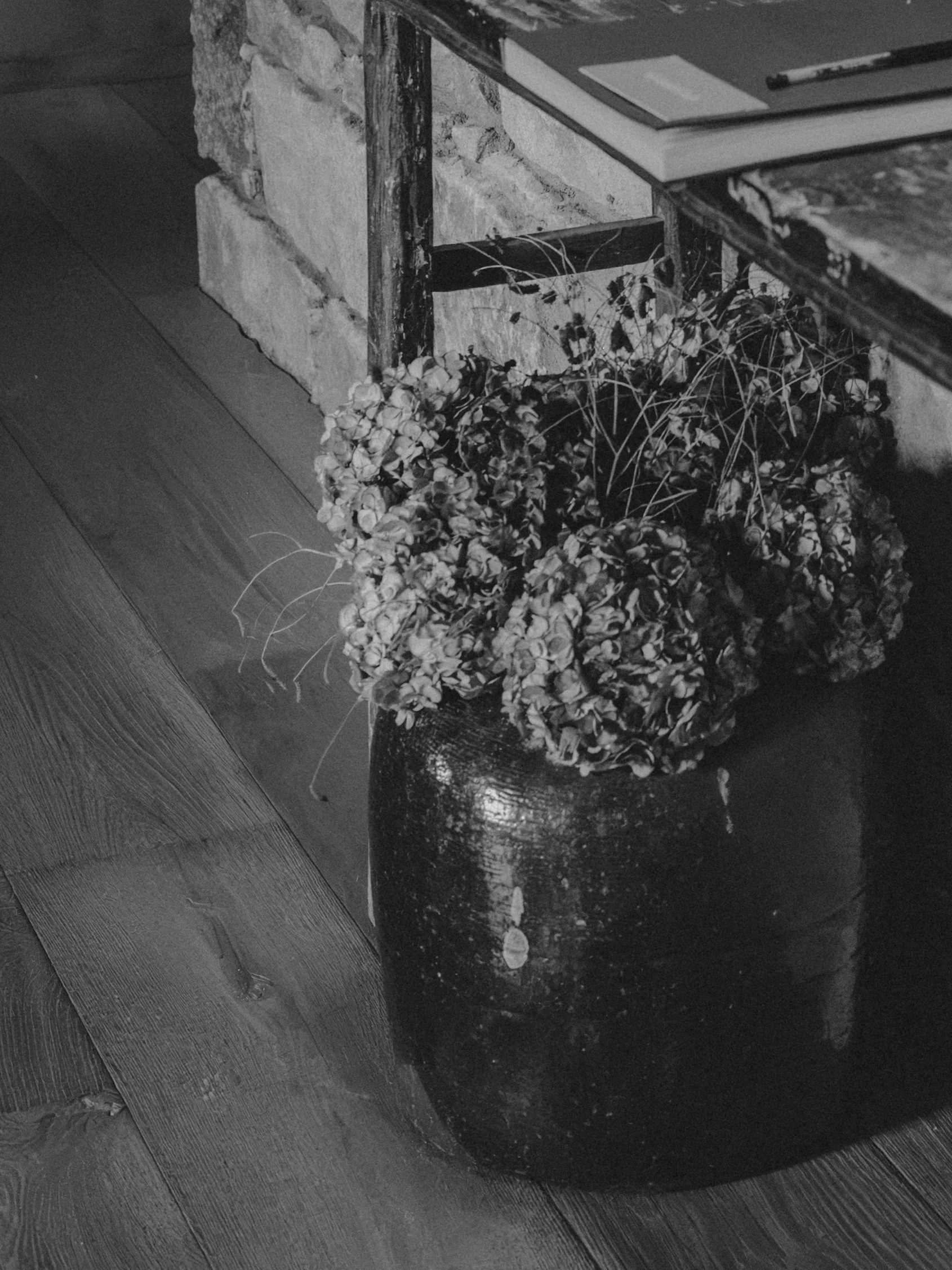 A black and white photograph of a tiled floor with a decorative planter holding a cluster of dried or wilted plant stems and leaves, placed against a rustic brick wall and a wooden table.