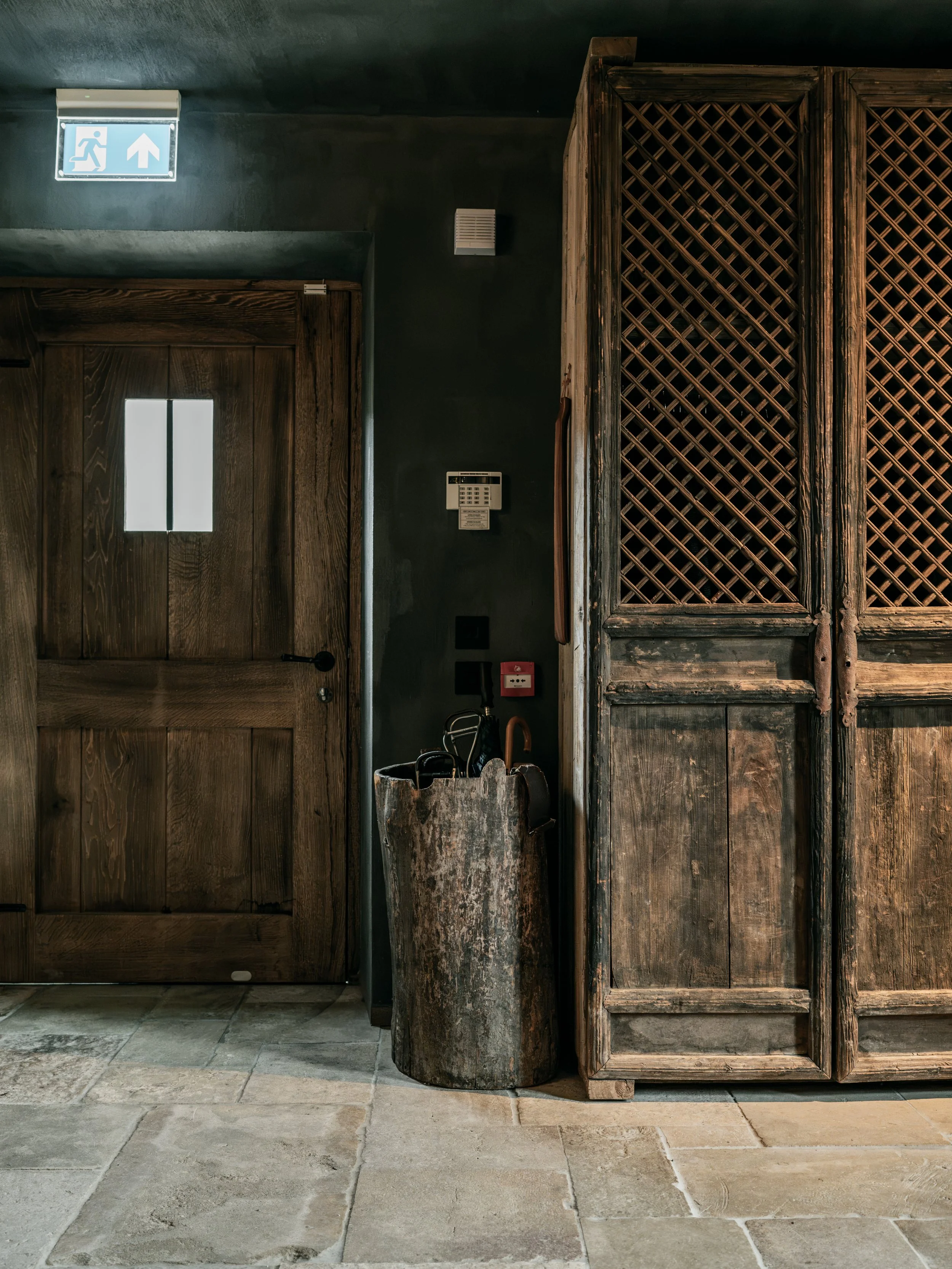 Interior of a rustic space with stone tile flooring, wooden door with vertical slats, a wooden cabinet with diamond lattice doors, a wall-mounted phone, an umbrella stand, and a ceiling-mounted emergency exit sign pointing upward.