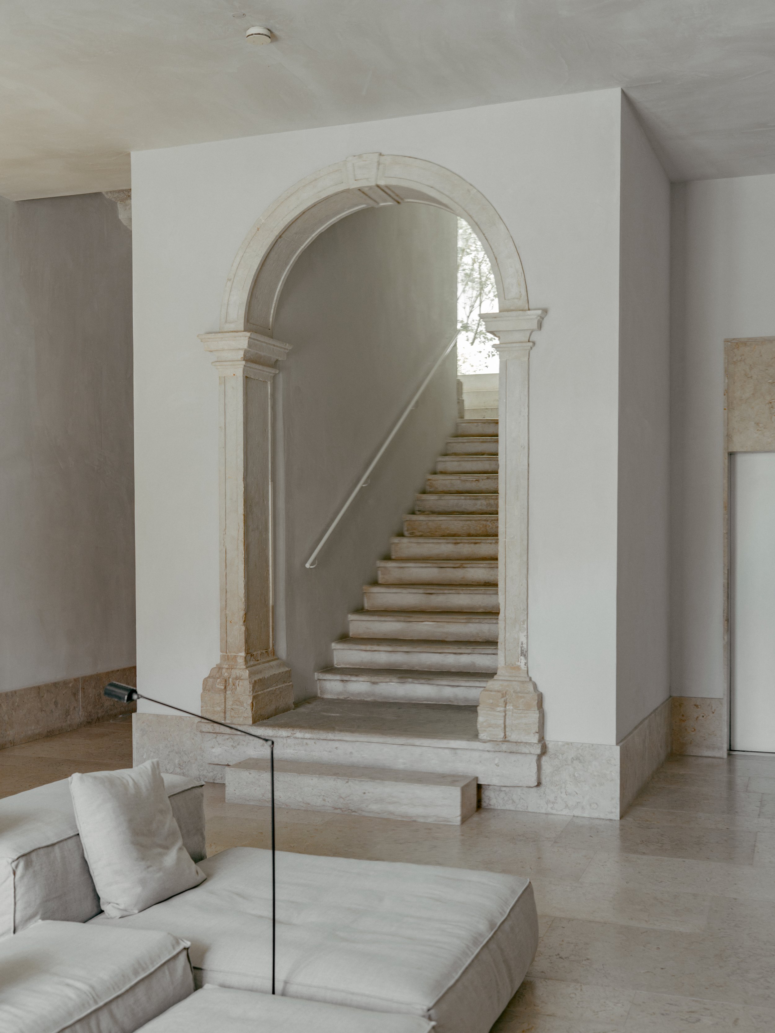 Interior view of a marble staircase framed by an archway with classical columns, leading up to a window with trees visible outside.