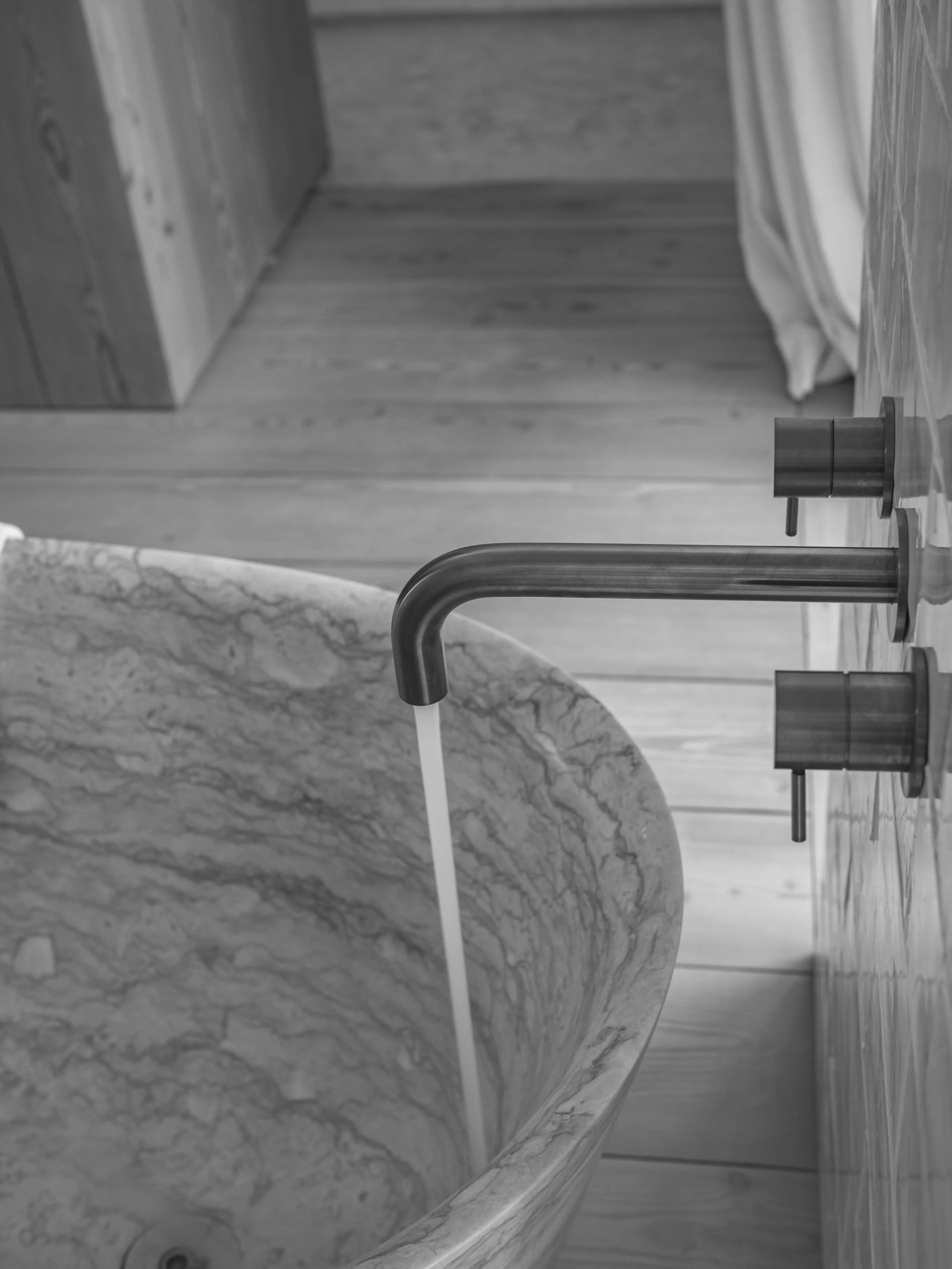 A marble bathroom sink with a silver faucet and water running, positioned in front of a wooden floor and surrounded by wooden and marble surfaces.