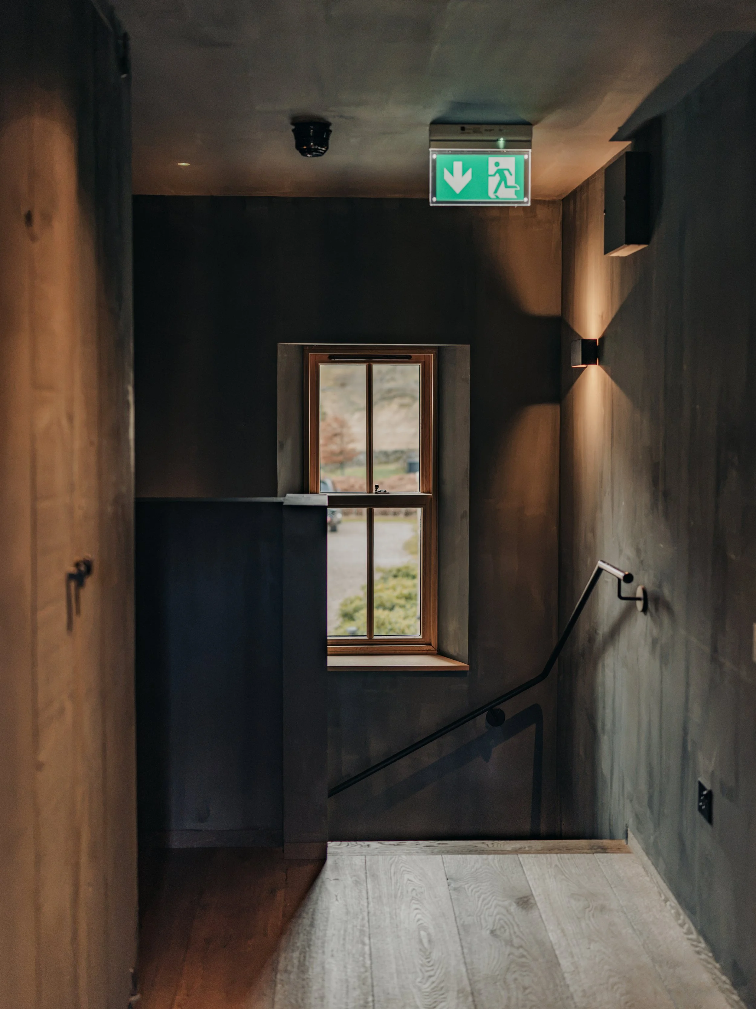A staircase enclosed by dark wood walls, with a window at the top showing an outdoor landscape. An emergency exit sign with a green background and white symbols is mounted on the ceiling, indicating downward direction and an exit.