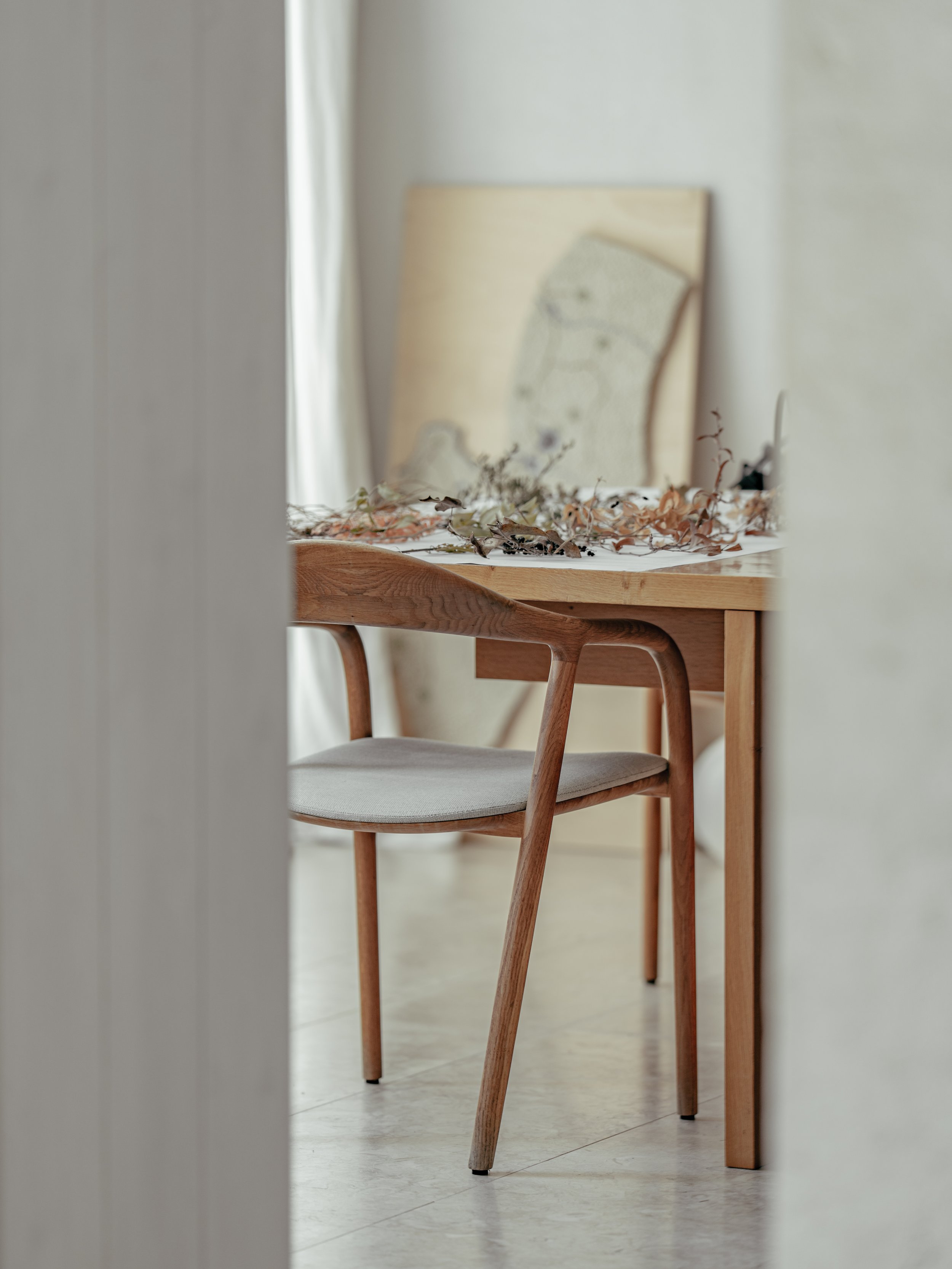 A wooden table with a printed map leaning against the wall, decorated with dried leaves, and a modern wooden chair with a cushioned seat, seen through a narrow gap between walls.