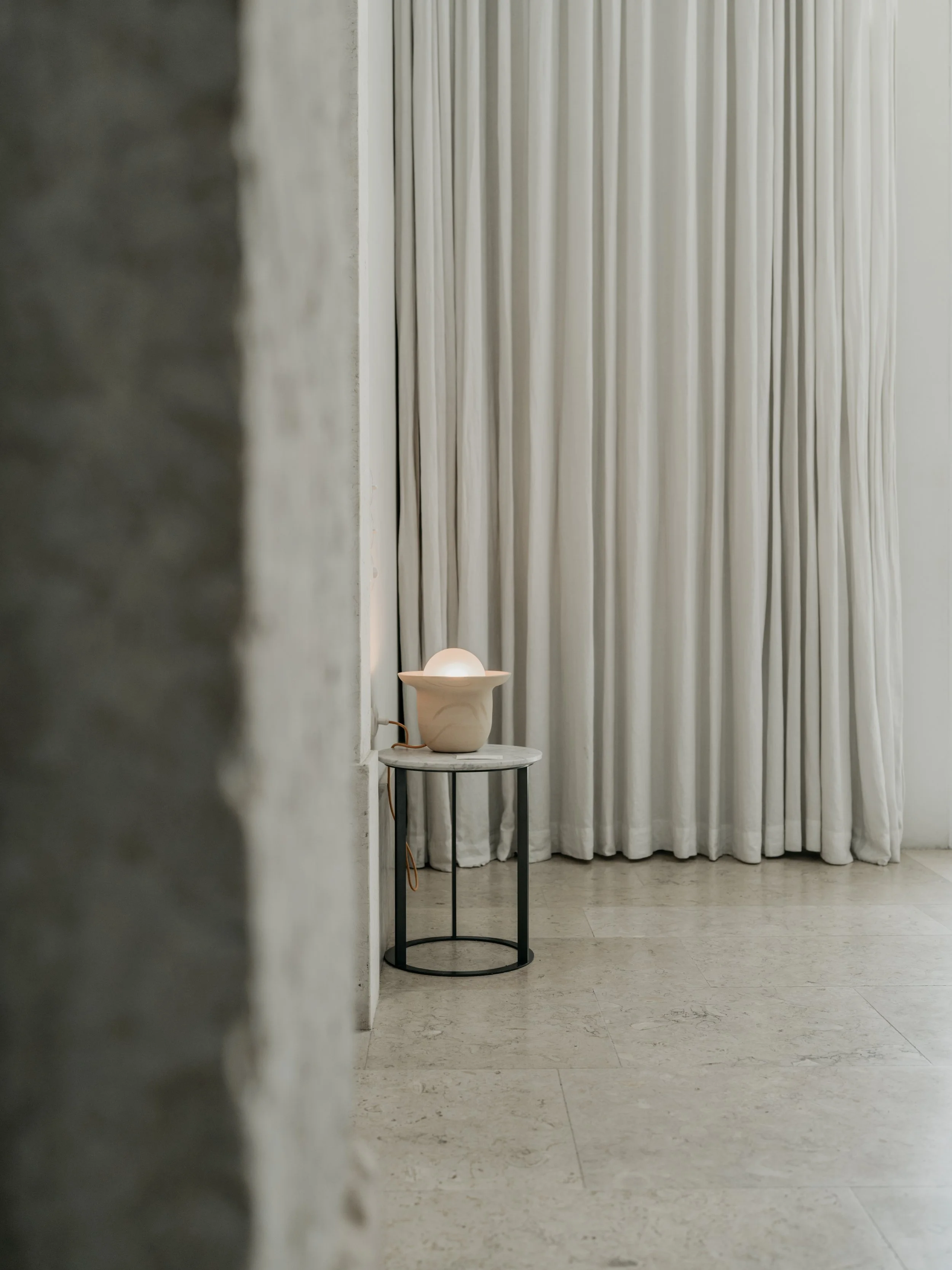 A view of a modern, minimalist room with a small round side table, a decorative lamp, and long white curtains covering the wall behind.