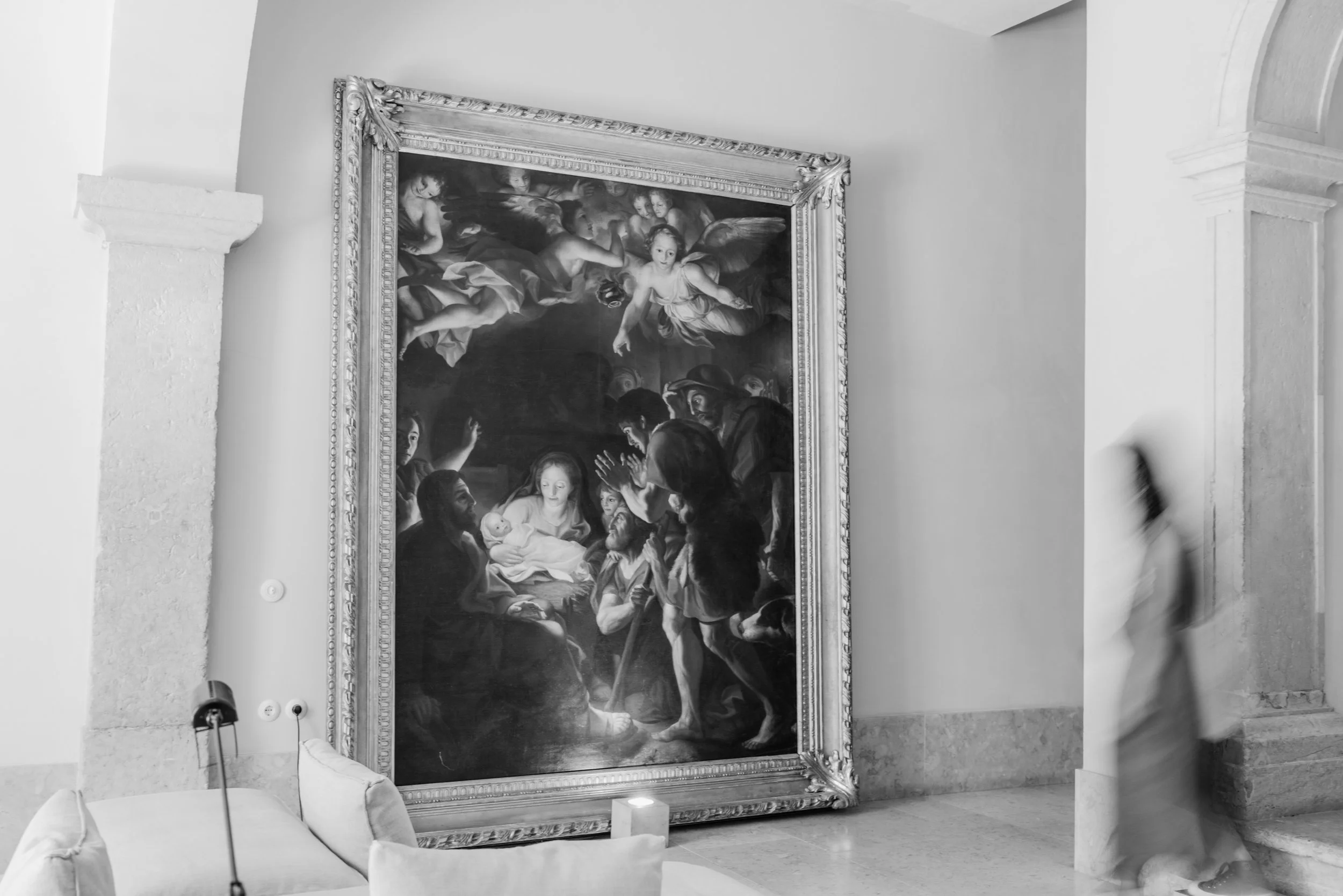 Black and white photograph of a classical religious painting displayed in a gallery, framed with ornate decorative molding, depicting a scene with angels and people, including a woman holding a baby.